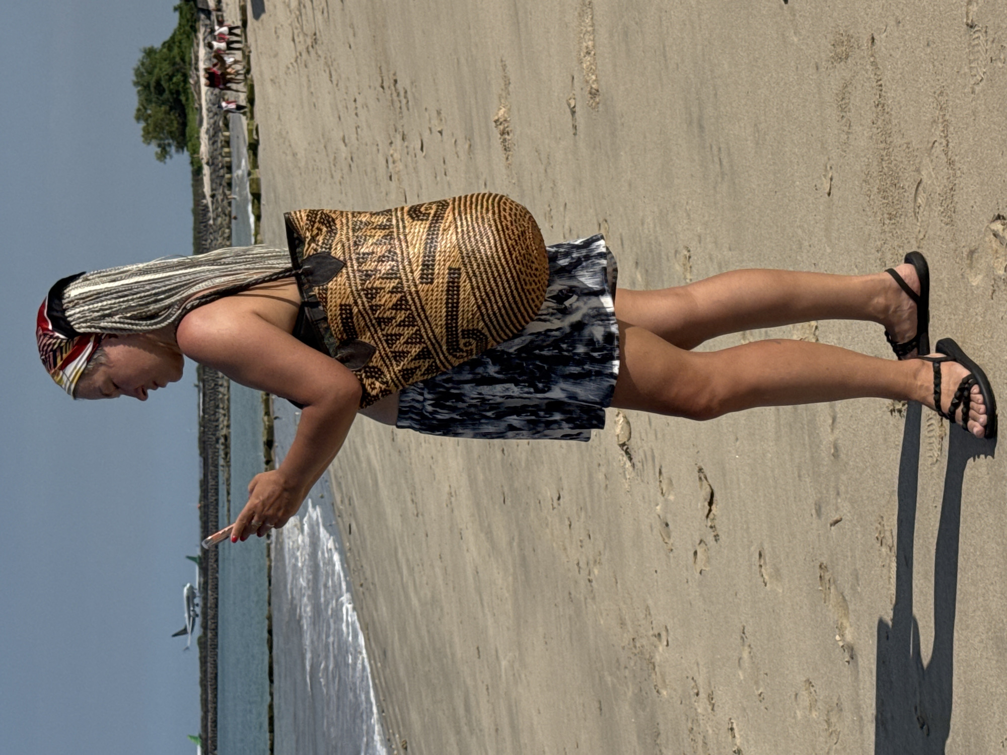 A woman stands on a sandy beach, angled toward the viewer's left, and is looking down at a cell phone in her hand. She is wearing a patterned headscarf, a large brown woven bag on her shoulder, and a black and white patterned dress. She has braided hair and is wearing black sandals. The beach appears to be wet near the water's edge, and footprints are visible in the sand. In the background, to the left, a breakwater is visible, and a plane is descending. Other people can be seen on what appears to be a walkway on the left of the image. Based on the descending plane and breakwater, it is reasonable to believe that this photo was taken in Kuta, Indonesia, near the airport.