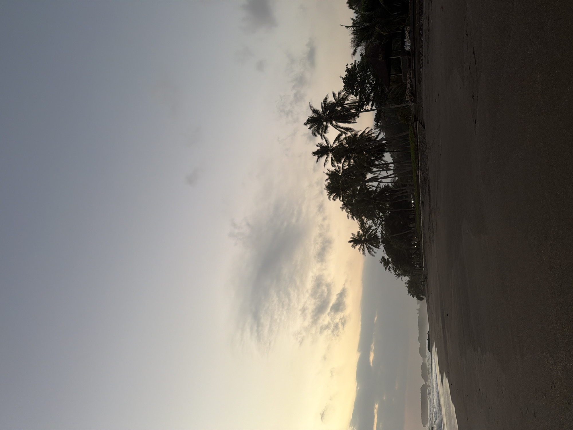 The image displays a wide-angle view of a dark sandy beach at dawn or dusk in Selemadeg Timur, Indonesia. The sky, occupying the upper two-thirds of the frame, exhibits a gradient from a pale yellow-orange at the horizon to a light blue-grey overhead, punctuated by scattered, wispy grey clouds. A singular, faint bright point, possibly a celestial body, is visible in the upper left portion of the sky. The ocean stretches along the left side, featuring small waves breaking onto the shore. In the far left distance, multiple dark rock formations or small islands are present in the water. A wide expanse of dark, wet sand covers the foreground and middle ground of the beach. Along the right side, a dense line of silhouetted palm trees and other tropical foliage forms a natural boundary. Within this tree line, a dark, thatched-roof structure, likely a gazebo or hut, is visible towards the right. Faint artificial lights are discernible behind the distant tree line. No individuals or prominent man-made objects are present on the immediate beach.