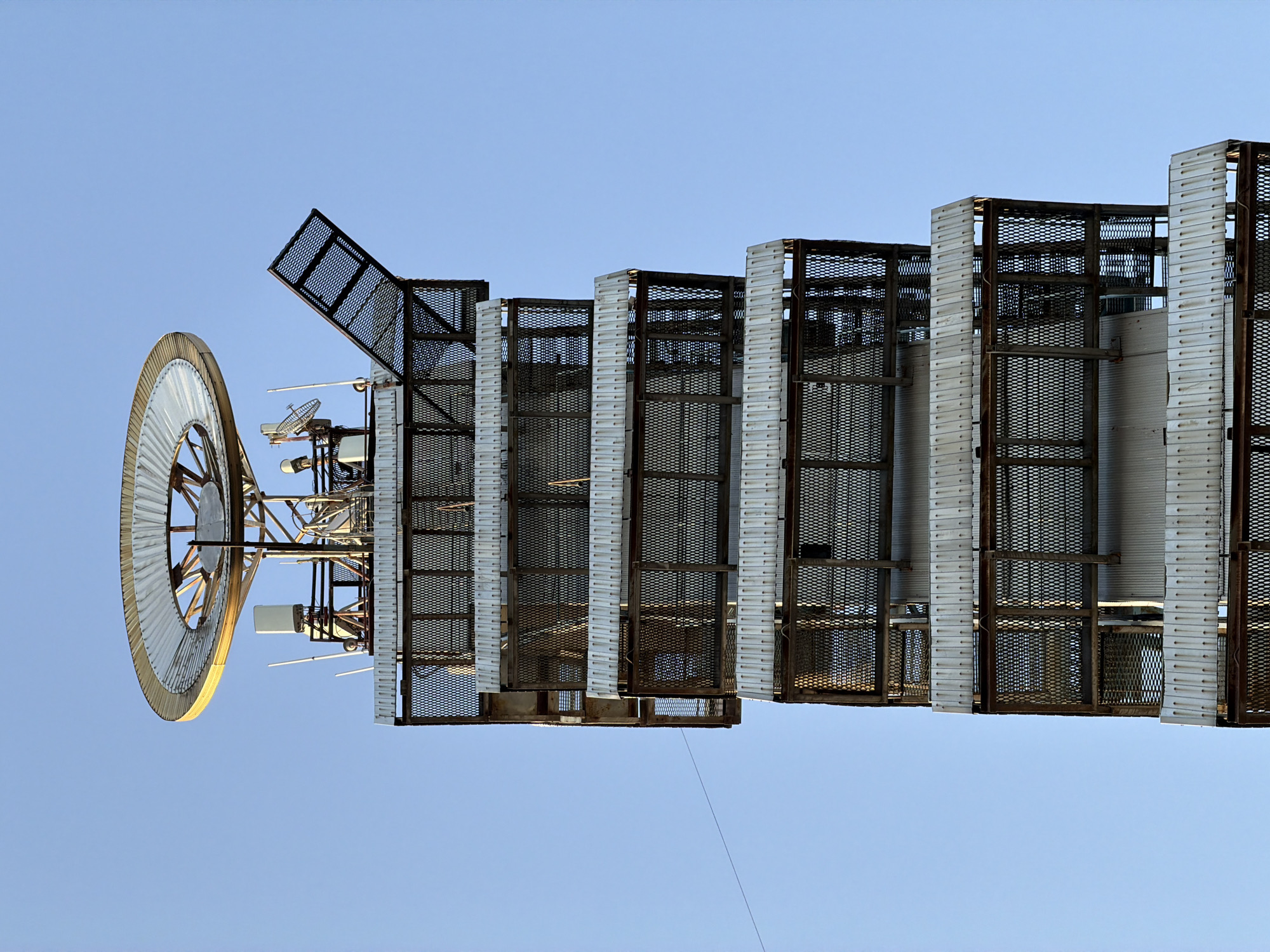 A close-up, low-angle, full-frame shot captures the upper portion of a tall, industrial tower against a clear, light blue sky. The tower has a segmented structure, with multiple levels featuring corrugated metal siding and caged platforms constructed from rusted metal grate. Several antennae and communication devices are visible near the top, along with a large, circular, metallic structure resembling a dish or radar. One of the caged platforms has an open, angled section. The overall impression is one of utilitarian infrastructure. The location is identified as the city of Hisarak, Uzbekistan.