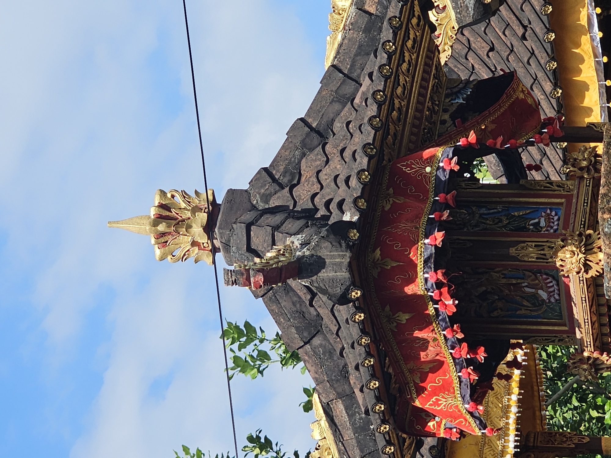 The image shows the roof of a structure with dark, tiled roofing and gold ornamentation. A red fabric with gold patterns is draped along the edge of the roof, adorned with red tassels. The structure appears to be a temple or shrine, possibly located in Denpasar, Indonesia, a city known for its Hindu temples. In the background, a blue sky with scattered clouds is visible, along with green foliage. A black wire runs horizontally across the frame. The overall scene suggests a religious or cultural site, possibly during a festival or ceremony given the decorative elements.