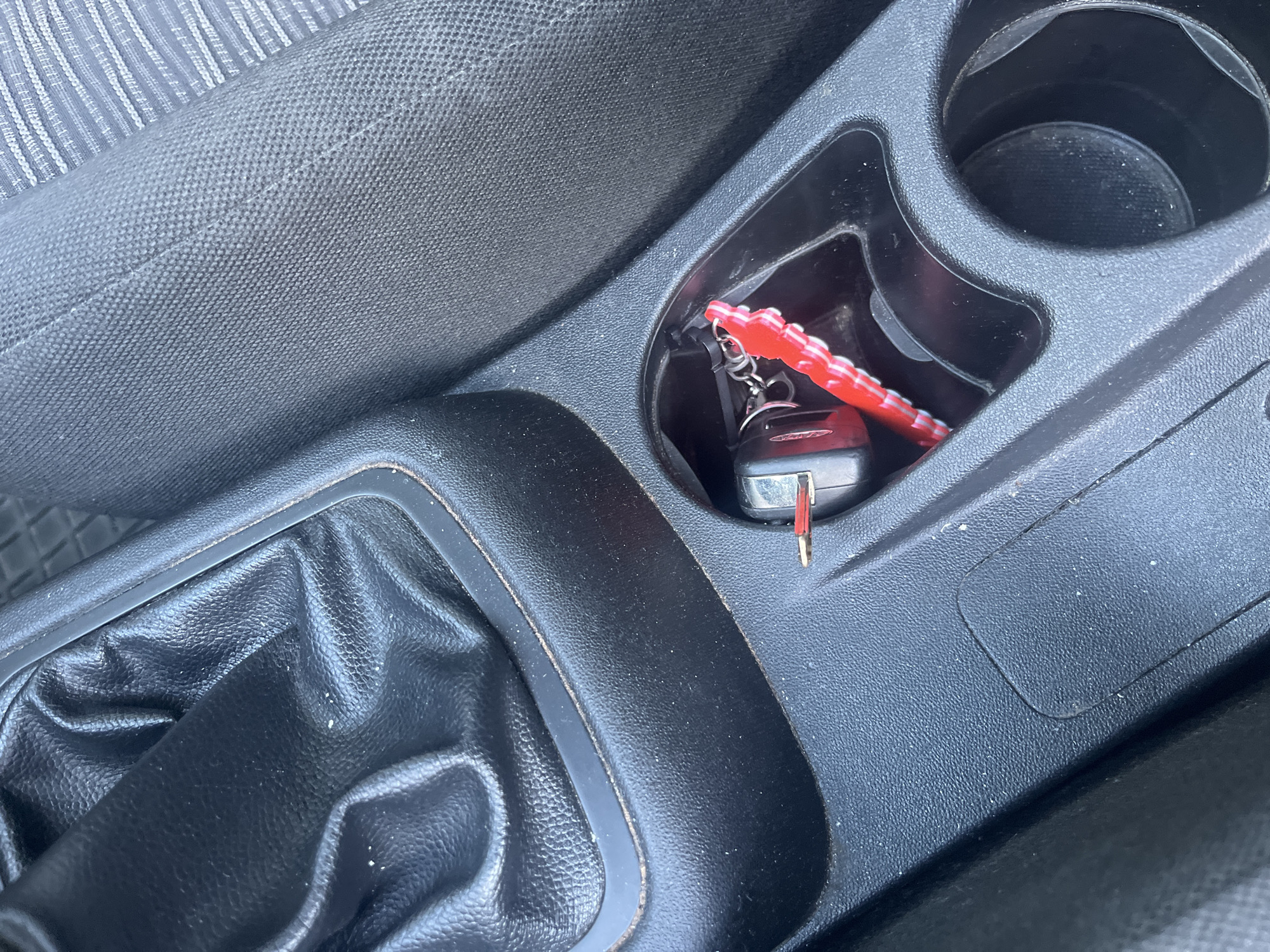The image shows the interior of a car, specifically the center console area. A car key with a red decorative keychain sits in the cupholder. A black gearshift lever is visible to the left of the cupholder. The car seat is partially visible on the left side of the frame. The scene takes place in a car, with no clear indication of the specific location in Blagoevgrad, Bulgaria. There are no people present in the image.