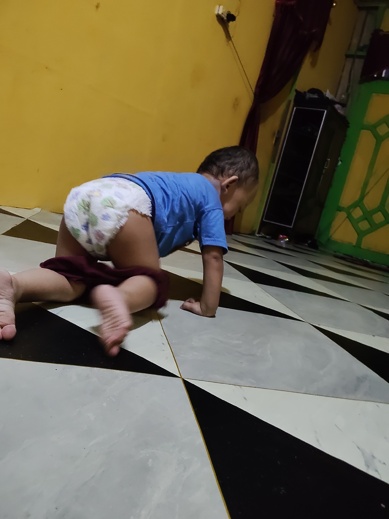 A baby is crawling on a checkered floor in a room with yellow walls. The baby is wearing a blue shirt and a diaper. A burgundy cloth is around the baby's legs. In the background, there is a black cabinet, a green window frame, and a red curtain. The location is a residential room in Tanah Grogot, Indonesia.