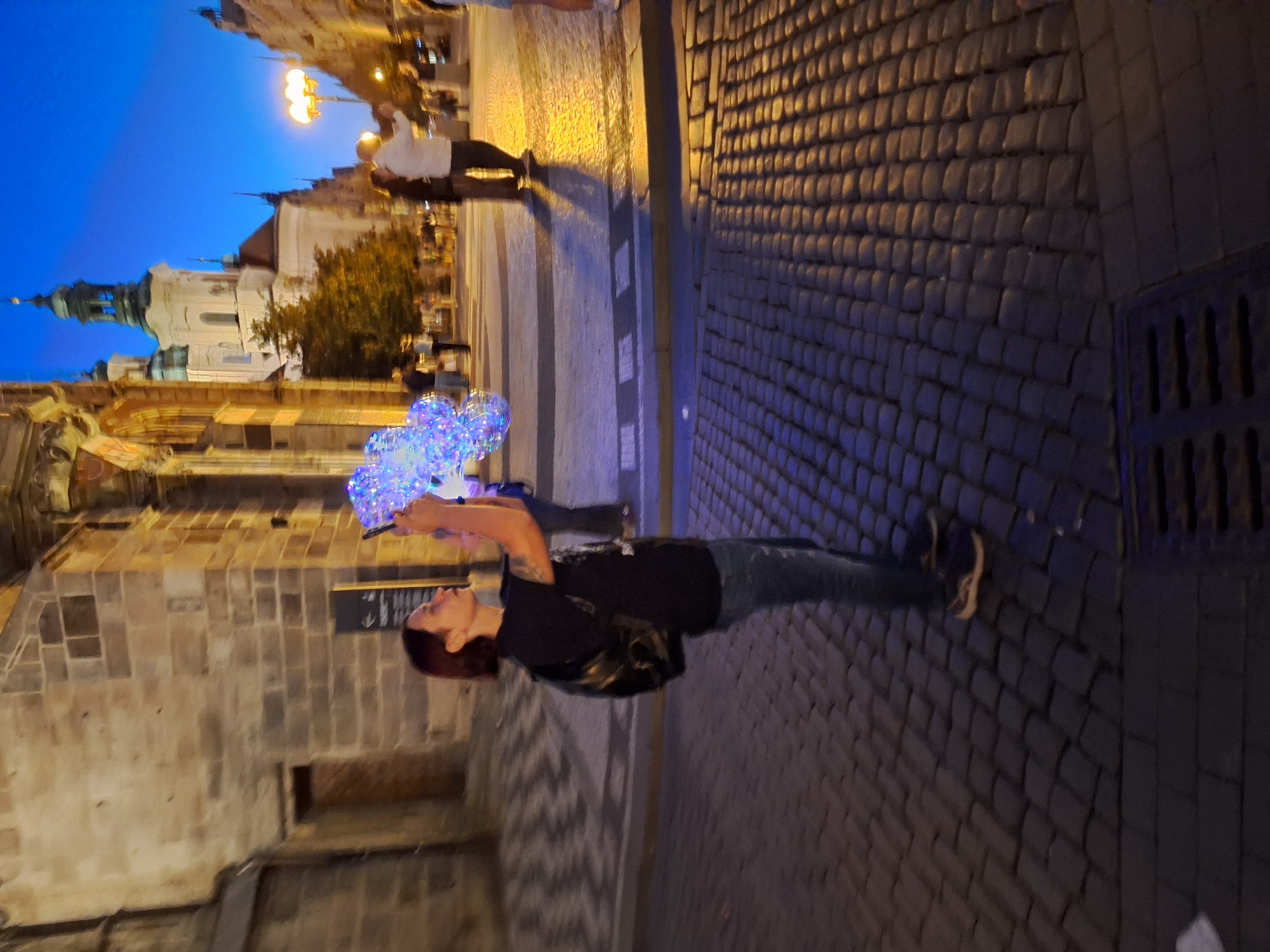 In Prague, Czechia, on a cobblestone street at dusk, a person with short, dark hair and a black backpack is holding a phone and a cluster of illuminated balloons. The balloons are a mix of blue and white lights, appearing to be made of a transparent material. The person is wearing a black t-shirt and blue jeans. Behind them, a building with stone brickwork and arched doorways is visible. In the background, other people are walking, and a church with two spires stands against a deep blue twilight sky. A streetlamp casts a warm glow on the cobblestones in the mid-ground.