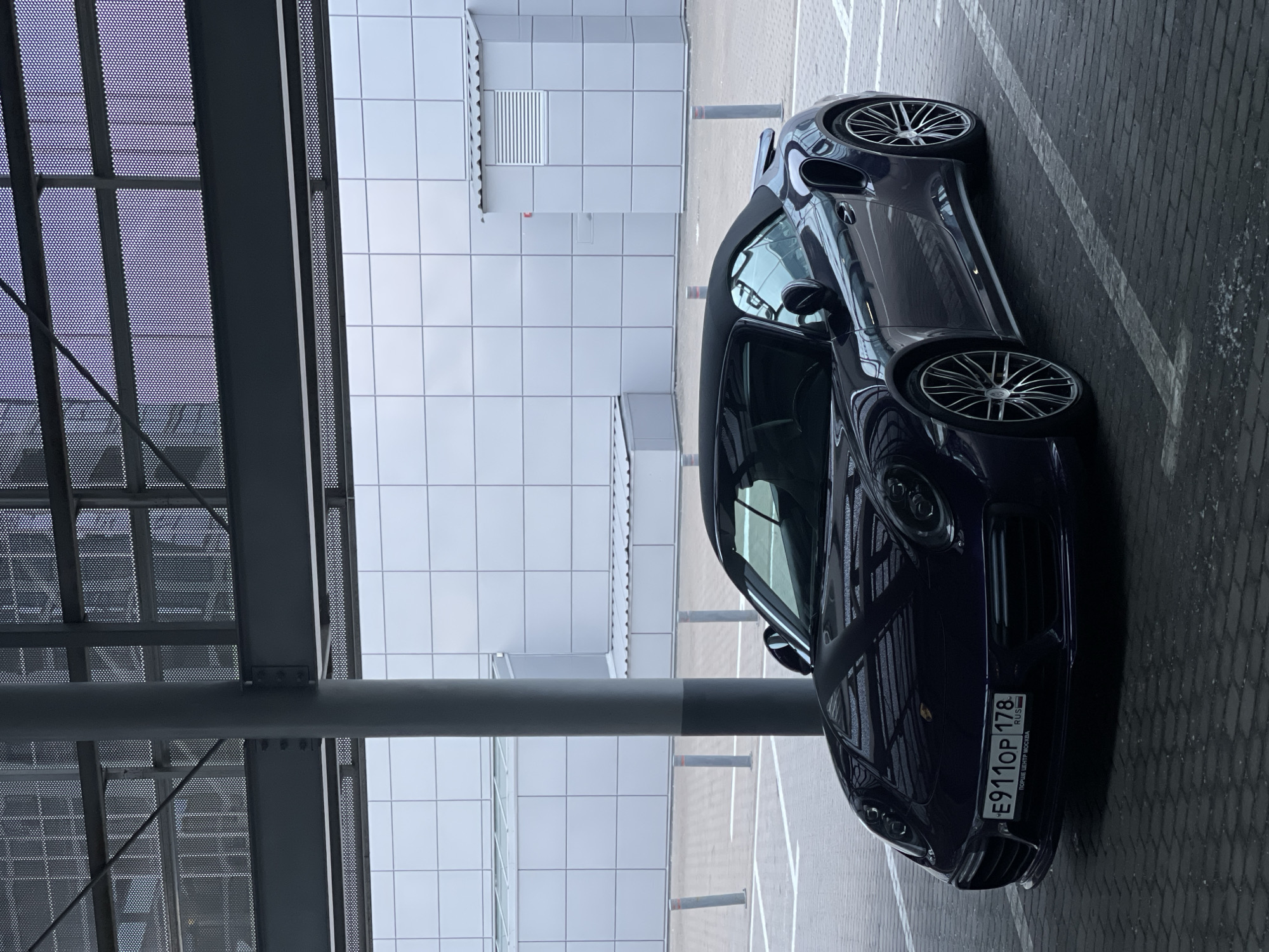 A dark purple convertible Porsche 911 Turbo S is parked in a lot. The car is facing left, with its front bumper and wheels clearly visible. The license plate reads "E911OP 178" with "RUS" below it, and "ПОРШЕ ЦЕНТР МОСКВА" on the bottom line. The car is parked under a modern, covered structure with a gray concrete pillar in the foreground. The background features a large building with a white tiled facade and a metallic roof structure overhead. The ground is paved with dark gray interlocking bricks. The scene appears to be an outdoor parking lot of a car dealership or service center in Moscow, Russia, given the text on the license plate. There are no people visible in the image.