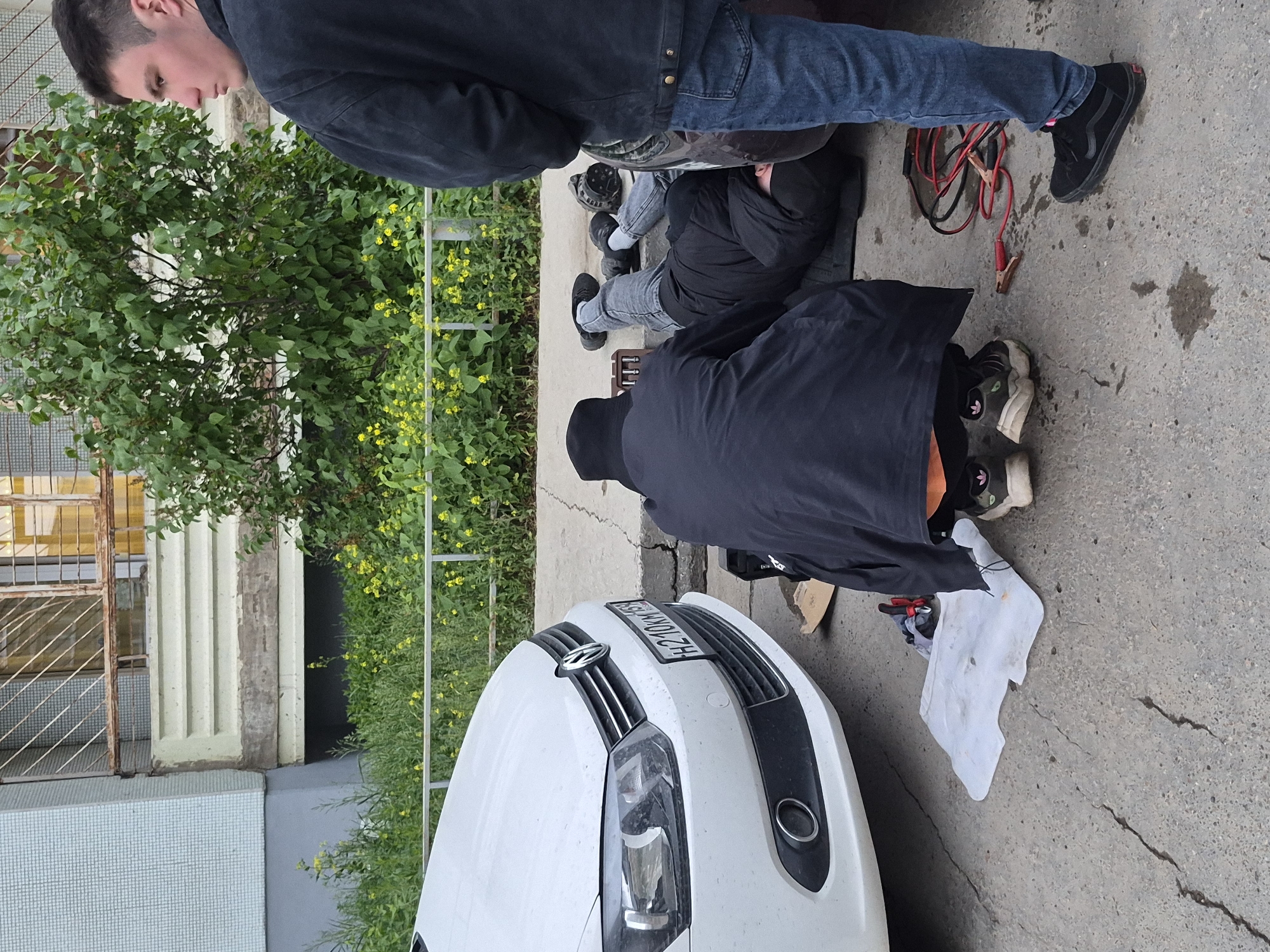 Outside, two people are working on a white car. One person is bent over the car, possibly working on the engine, while the other person is standing nearby. Jumper cables, tools, and a white rag are on the ground. The location appears to be Chaikovsky, Russia, based on the license plate. A building with a fenced window is visible in the background. Green foliage is also present.