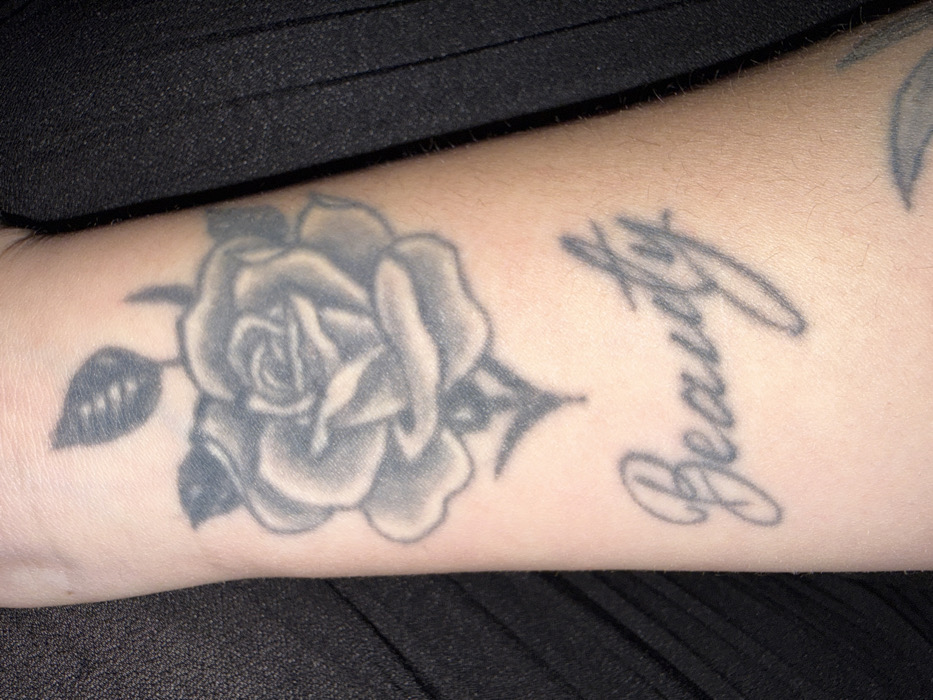 The image displays a close-up, slightly angled view of a human forearm. A prominent grayscale tattoo of a rose, complete with leaves and a stem, is situated towards the upper section of the arm. Directly beneath the rose, the word "Beauty" is tattooed in a decorative script font, also in grayscale. A partial, indistinct grayscale tattoo is visible at the bottom right periphery of the forearm. The skin of the arm is light-toned and exhibits fine hair. The background consists of a dark, textured fabric, likely clothing, on both sides of the arm. The scene is located in Polski Izvor, Bulgaria.