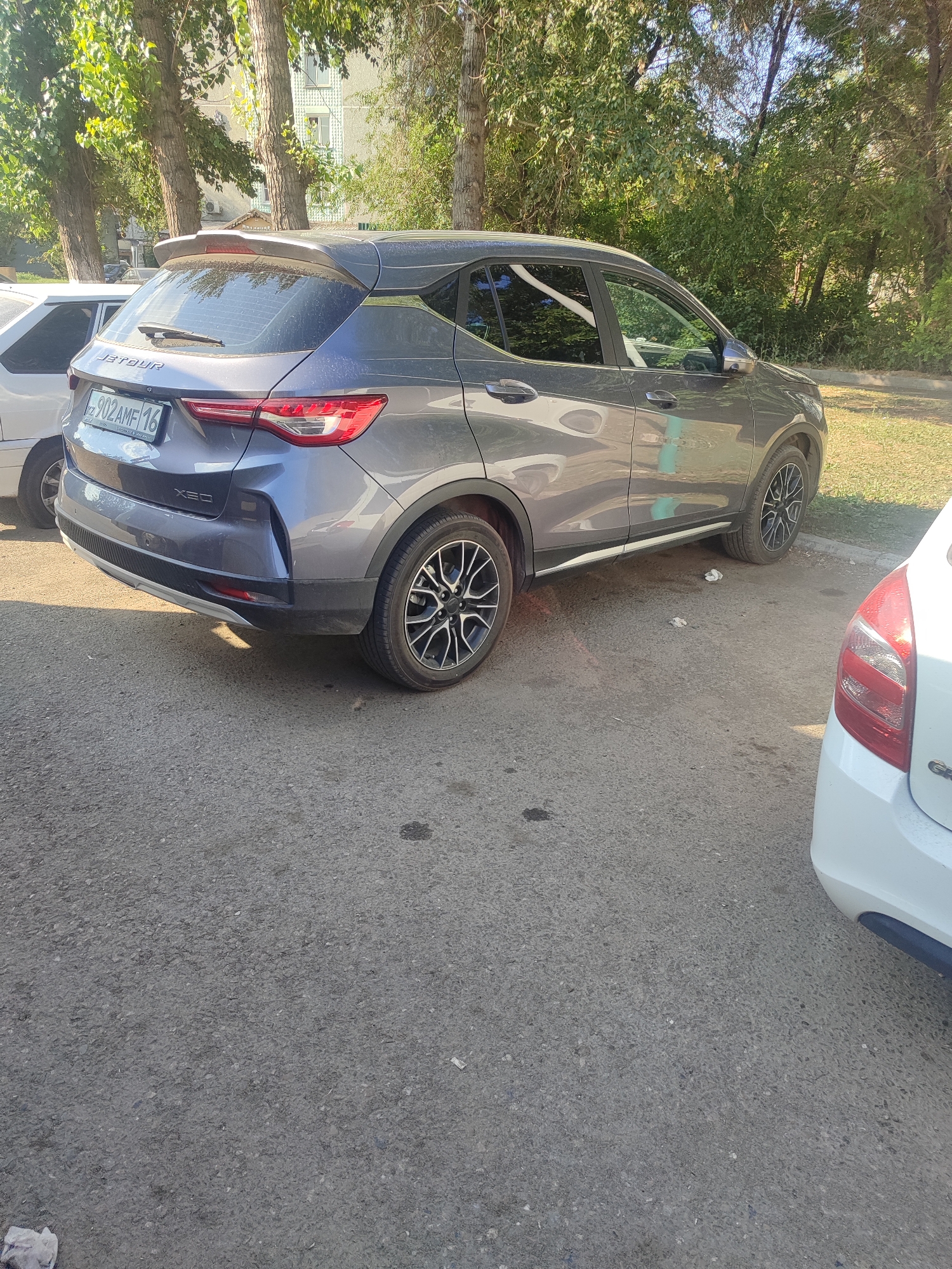 The image shows the rear and side of a gray Jetour X30 SUV parked on an asphalt surface in Oskemen, Kazakhstan. The car has a license plate that reads "KZ 902 AMF 16". A white car is partially visible in the foreground to the right. In the background, there are trees, foliage, and a building. The scene appears to be a street or a parking area, possibly in a residential or commercial district. The lighting suggests it is daytime. There are no people visible in the image.