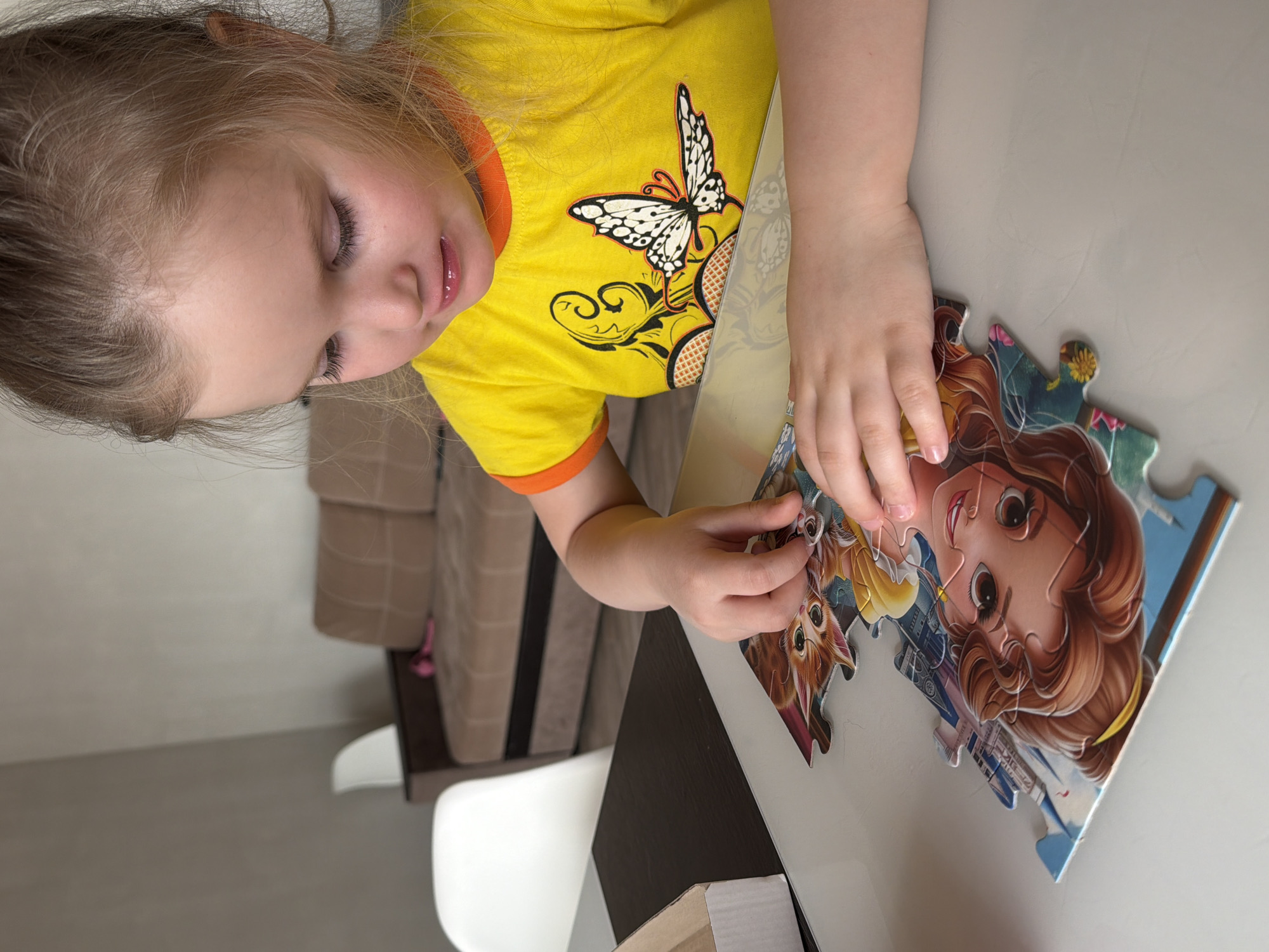 A young girl with blonde hair, wearing a yellow shirt with an orange collar and a butterfly graphic, is sitting at a table and putting together a puzzle. The puzzle features a cartoon image of a girl with brown hair and a cartoon cat. The scene is indoors, with a sofa and a chair visible in the background. The location may be Chaikovsky, Russia, although this is not visually verifiable.