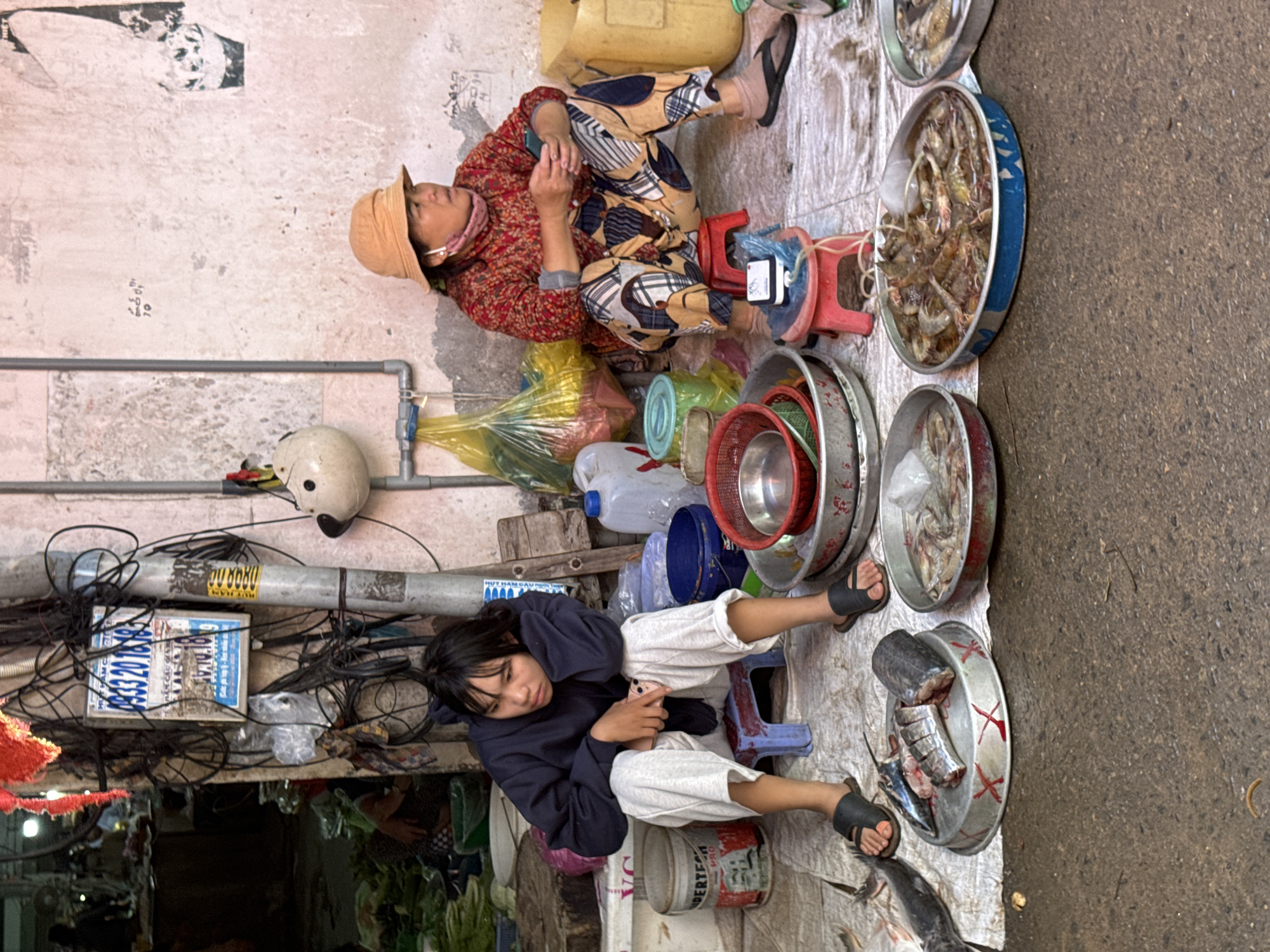 This image depicts a street scene in Mũi Né, Vietnam, where two women are seen sitting amidst a market stall. On the left, a young woman, wearing a dark blue hoodie and light grey pants, is seated on a small blue stool. Her legs are crossed, and she is holding a smartphone. She is wearing black flip-flops. To her right, an older woman, dressed in a vibrant red and black patterned shirt and similarly patterned pants, sits on a red stool. She wears a beige sunhat and a pink neck scarf, and also has black flip-flops on. Both women appear to be waiting or resting.
The environment is a bustling marketplace, indicated by the presence of various items for sale. In front of the women are several large metal bowls containing seafood. One bowl holds cut fish, and others contain shrimp. To the left of the young woman, a large bucket labeled "PERTECN PRO" is visible. Behind the women, there are yellow plastic bags filled with goods and a white water cooler. A pole covered in tangled electrical wires and a sign with Vietnamese text stands prominently behind the young woman. The ground is paved with concrete and appears wet in some areas, suggesting a recent cleaning or rain. The background shows more market stalls and a textured, somewhat dilapidated wall. The overall impression is one of everyday commerce in a Vietnamese coastal town.