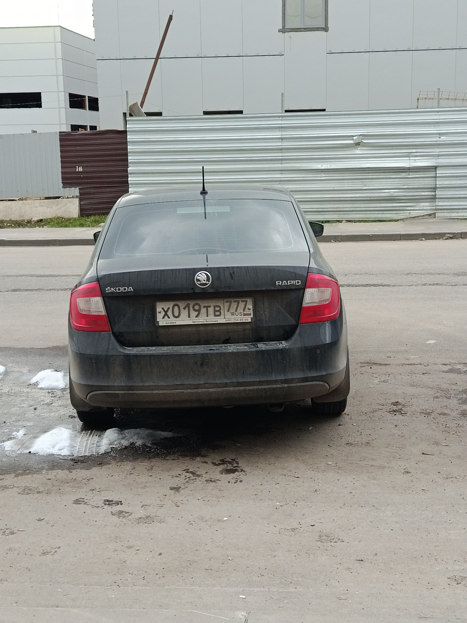 The image shows the rear of a black Škoda Rapid parked on an asphalt surface with scattered patches of melting snow and debris. The license plate reads "X019TB777" with the Russian flag emblem, indicating a vehicle registered in Russia. The license plate includes the text "RUS" and a phone number beginning with (495). The vehicle is facing the camera, with a corrugated metal fence and a multi-story building visible in the background. The scene appears to be an urban environment, possibly a street or parking area near a construction site or industrial area. Given that the vehicle's registration code is 777, this suggests the location might be within the city of Moscow, Russia. There are no people visible in the frame.