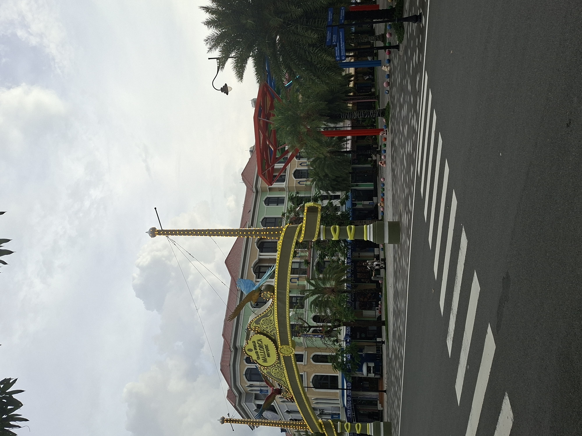 The image depicts the entrance archway of "Grand World Phu Quoc" in Phu Quoc, Vietnam, under an overcast sky. The archway is adorned with yellow lights and features two large, colorful macaws on either side. Behind the archway, a multi-story building with a red-tiled roof and pastel-colored facades stands. Several palm trees are scattered throughout the scene, some with string lights. In the foreground, a pedestrian crossing is marked on an asphalt road. The area in front of the building appears to be a public square decorated with various colorful balls and some small figures. A single person is visible sitting on a bench near the building. The overall impression is that of a commercial or entertainment district, possibly a theme park or resort.