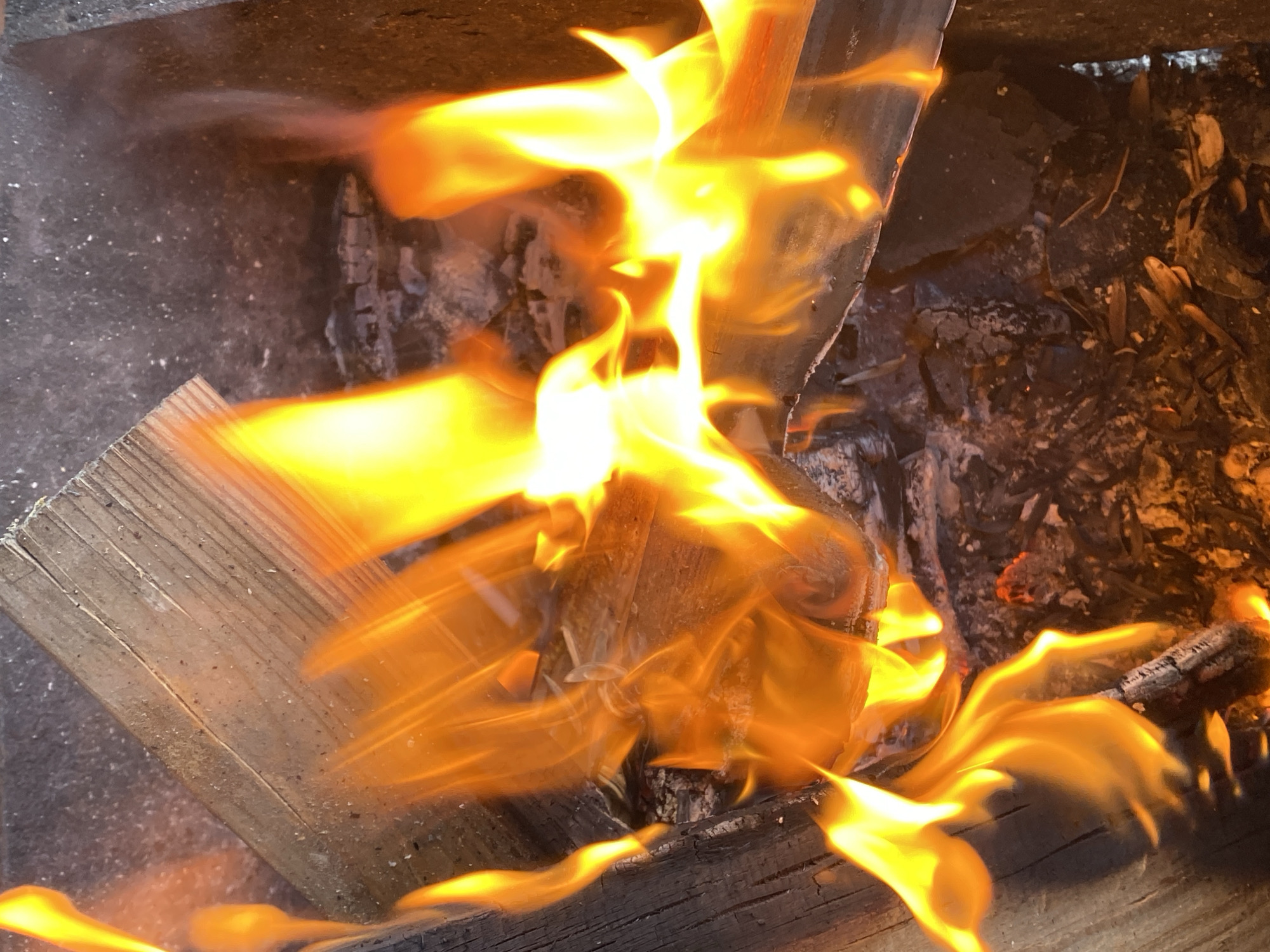 A close-up captures burning wood in a fire pit. Flames engulf the wood, consuming it. Ash and charred remains surround the burning logs. The fire pit is located outdoors, likely in a residential or recreational area. There is no information to infer that this image was taken in the city of Chaikovsky, Russia. There are no people visible in the image.