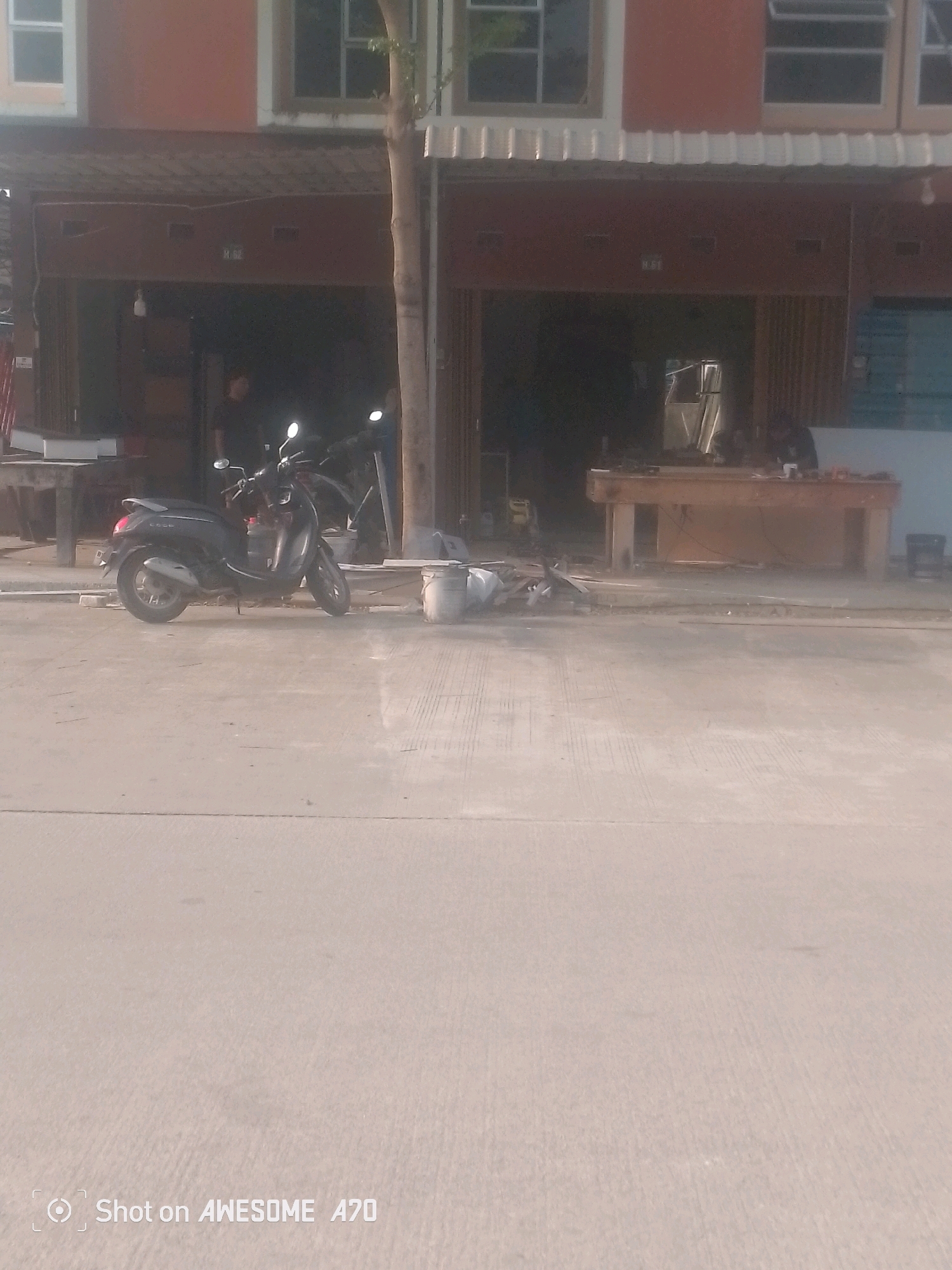 A dark-colored scooter is parked on a concrete road in front of a building. The building has multiple stories with orange and white facades and windows. There appears to be a shop or business on the ground floor with a wooden counter visible inside. The scooter is the primary subject in the foreground, and there is a tree to the right of the scooter. The scene is outdoors, with a clear sky, and appears to be a street in a city.
