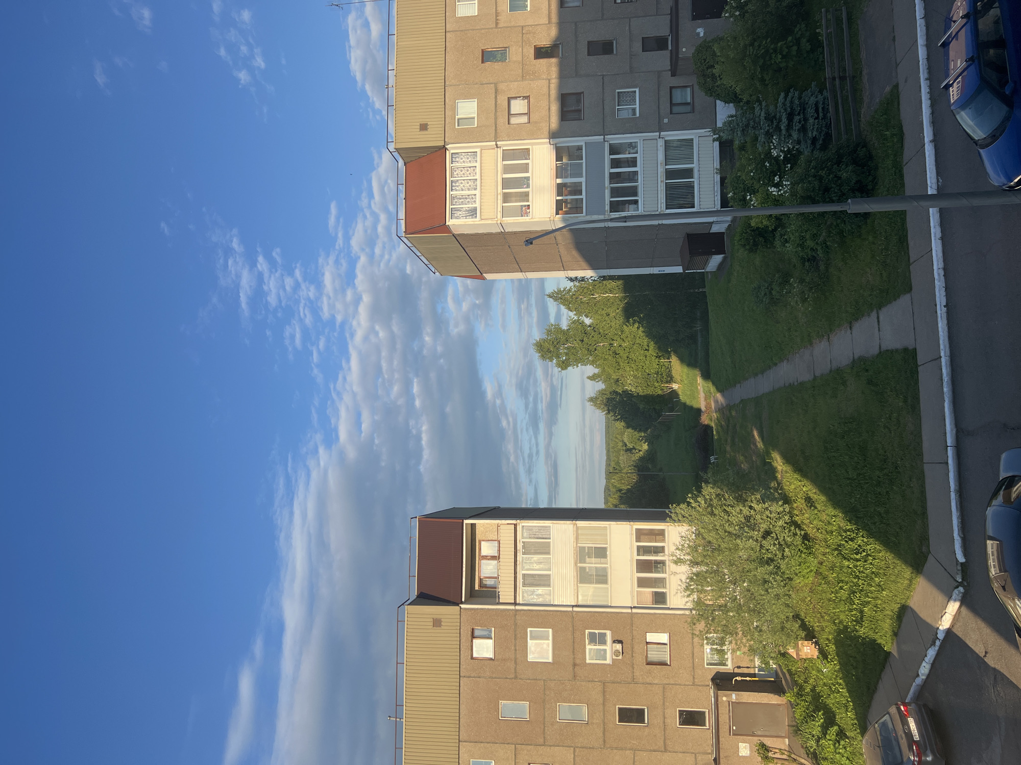 The image captures a residential scene in Марковский, Russia, featuring two multi-story apartment buildings under a bright, partly cloudy sky. The photograph is taken during the late afternoon or early evening, characterized by the low sun casting long, distinct shadows across the landscape.
In the foreground, on the left, stands a concrete panel apartment building with numerous windows and several enclosed balconies or loggias. The building has a multi-toned roof, part dark brown and part lighter grey. On the ground level near this building, a dark grey or black car is parked, with a visible Russian license plate displaying "Е408РО" followed by a partial region code starting with "1".
Further back and to the right, a second, similar apartment building is visible. This building also features the characteristic concrete panel construction and enclosed balconies, with parts of its facade obscured by shadows. An antenna is noticeable on its roof.
Between and in front of the buildings, there is an expansive green area composed of grass, various trees, and bushes, some of which are flowering white. A paved pathway meanders through this green space, with sections illuminated by direct sunlight and others covered by the long shadows from the buildings and trees. On the far right of the image, an asphalt road or parking area is visible, where a blue car is partially parked. A lamppost or utility pole stands prominently in front of the blue car.
The sky occupies a significant portion of the upper image, a clear, vibrant blue dotted with scattered, fluffy white clouds, indicating pleasant weather. The overall impression is a tranquil and typical urban residential setting during a sunny day.