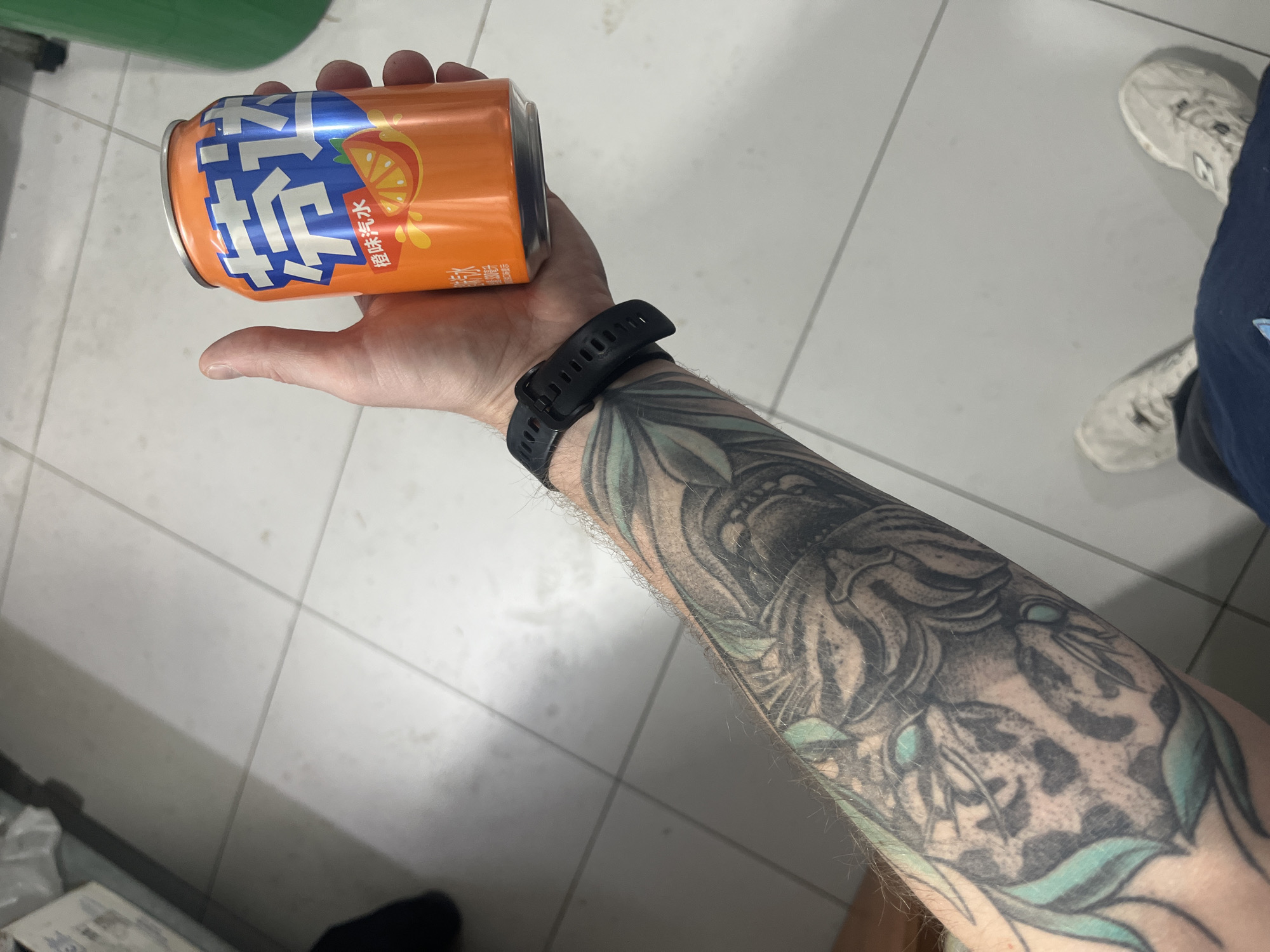 A person's left arm, adorned with a tattoo of a tiger and green leaves, is visible in the foreground. The arm is holding an orange soda can with Chinese characters and an image of an orange slice. The can is labeled "带汽" and "橙味汽水". The person is wearing a black wristband and a dark blue shirt. The background shows a tiled floor, characteristic of indoor spaces, and out of focus, a green object and white packaging. The scene is indoors, possibly a store or a home, and not in the city of Yablonovskiy, Russia.