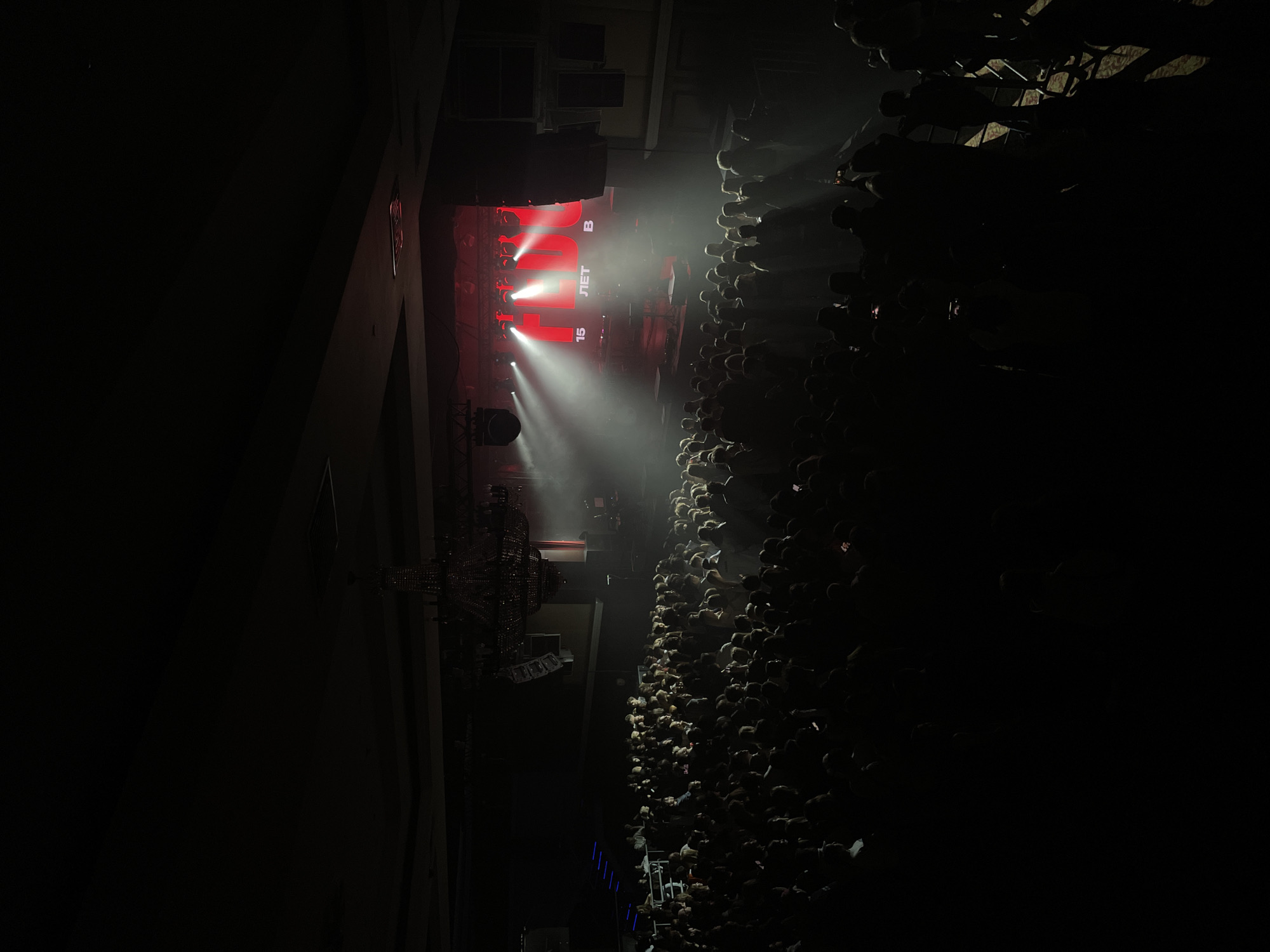 This high-angle shot captures an energetic indoor concert or live event in Kazan, Russia. The image is oriented to show a large crowd facing a brightly lit stage in a dark venue.
The scene is dominated by strong stage lighting, with multiple beams of white and red light cutting through what appears to be atmospheric haze or smoke. These lights illuminate parts of the stage and the audience closest to it. On the stage, a large screen or banner is visible, displaying text in what appears to be a Russian context. Clearly visible are the large number "15" and the Russian word "ЛЕТ" (LET), meaning "years." Above this, the letters "ЛЮБ" (LYUB) are discernible, possibly part of a larger word or phrase. Another word, "JET" or "RET," is spelled out in white capital letters on the screen, potentially indicating a band or event name. A single letter "B" is also visible on the right side of the stage display. A red "EXIT" sign is visible on a wall to the left of the stage.
Hundreds of attendees fill the floor, standing shoulder-to-shoulder and facing the stage. Their heads and shoulders form a dense mass, mostly silhouetted against the bright lights. Many individuals in the crowd are seen holding up their smartphones, suggesting they are recording or taking photos of the performance.
The venue's architecture is visible, with dark walls and a large, ornate dark chandelier or lighting fixture hanging on the left side. The overall ambiance is dark and immersive, characteristic of a nighttime concert or performance, focusing all attention on the illuminated stage and the activity unfolding there.