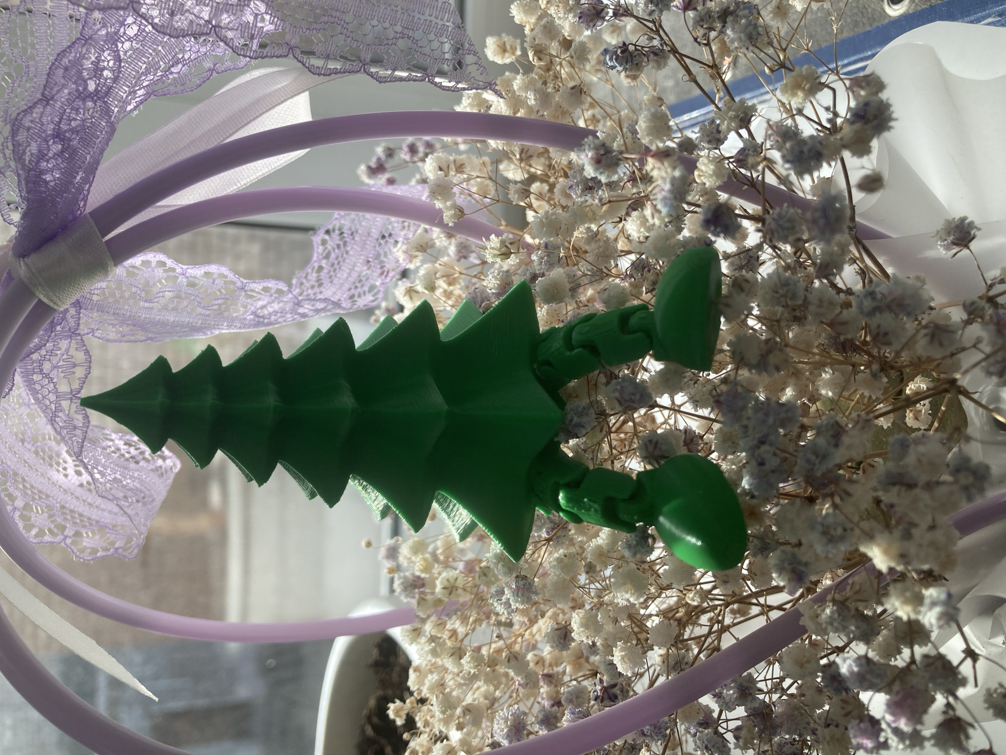 The image captures an indoor still-life arrangement, centrally featuring a bright green, 3D-printed articulated toy. This toy, resembling either a stylized Christmas tree or a creature with a serrated, tree-like spine, lies horizontally across the frame. At one end, it has two rounded, bulbous appendages, suggesting feet or hands.
To the left of the green object, a decorative arrangement is visible. This includes several thin, curved, light purple plastic tubes or wires, possibly part of a headband or similar accessory. Intertwined with these tubes is delicate, light purple lace fabric, along with a narrow white ribbon or band.
Filling the right side and background of the image is a substantial cluster of dried baby's breath (Gypsophila) flowers. These small, airy flowers are predominantly white, with subtle hints of faded purplish or pale blue in some of the flower clusters.
The scene is illuminated by soft, diffused natural light, suggesting it is daytime and possibly near a window. A blurred white object, potentially a plant pot or a window sill, is visible at the bottom of the frame. There are no people, visible text, or discernible activities taking place. The setting is an indoor space.