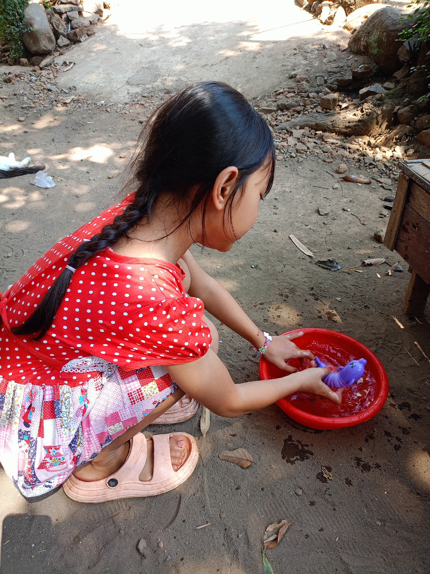An outdoor, eye-level medium shot shows a young girl playing with a purple toy in a red bowl of water. She is sitting on the ground, facing right. She wears a red, polka-dotted dress with ruffled trim on the skirt and light-colored sandals. Her black hair is braided into a single plait. The ground is dirt and gravel, with scattered leaves and rocks. In the background, a stone wall and large rocks are visible. A wooden structure is on the right edge of the frame. The scene appears to be in a rural area.