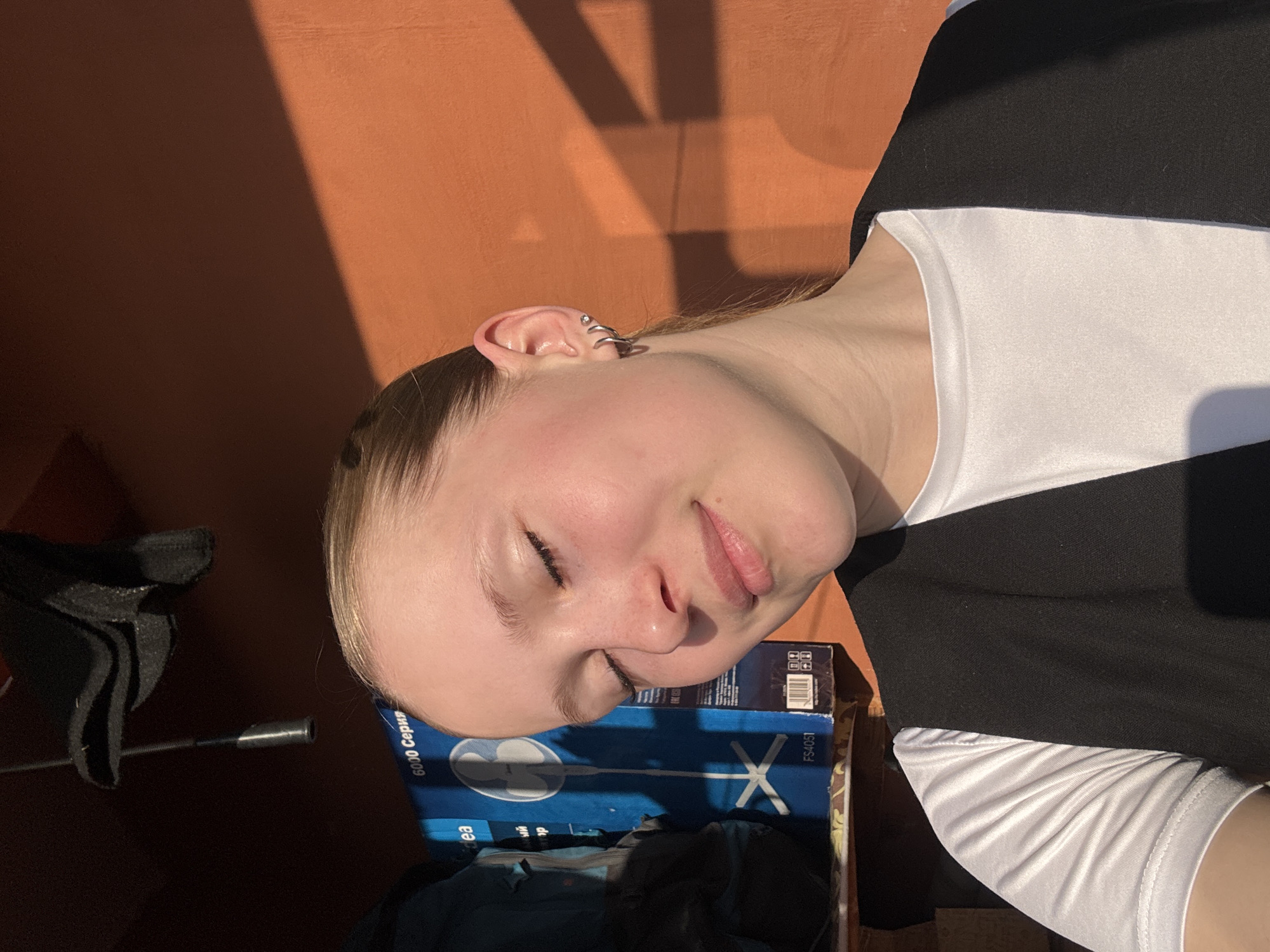 A young woman with fair skin and blonde hair is lying on a box of a fan with a logo and the word "idea" written on it and the number FS4051. She is wearing a black and white striped top. Her head is turned to the side and she is lying on a box. There is a microphone in the background. The background includes an orange wall with shadows. The picture appears to have been taken in Chaikovsky, Russia.
