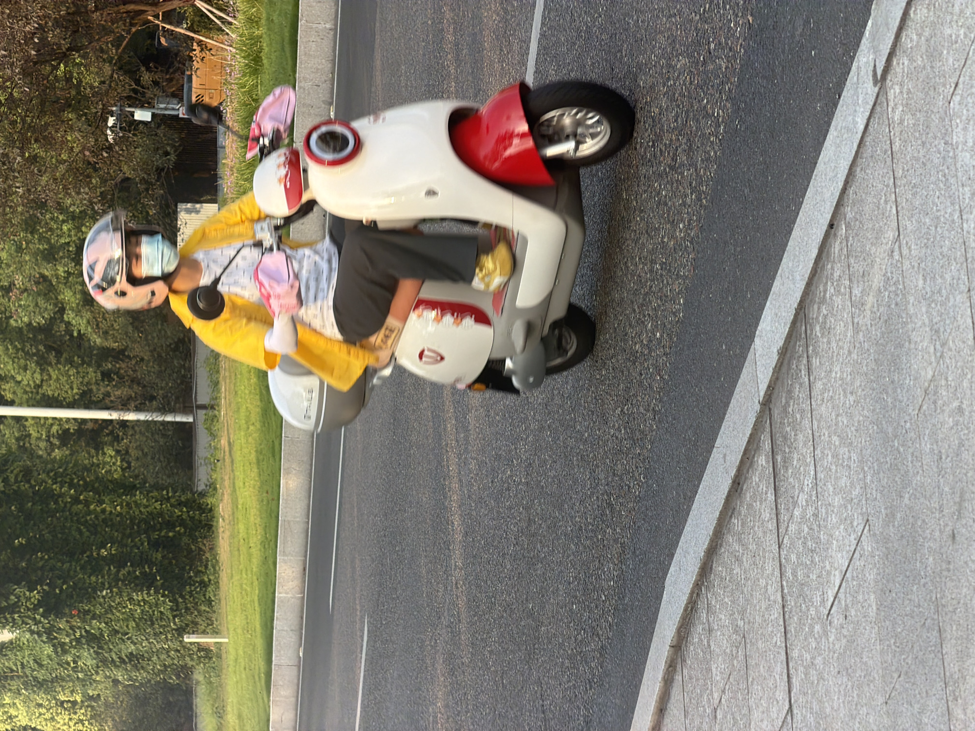 A person wearing a yellow jacket, a light blue face mask, a pink helmet with a clear visor, and light pink arm sleeves rides a white and red scooter on an asphalt road. The scooter has a white body with red accents on the front fender and side panel, and a red ring around the headlight. The person's legs are clad in dark gray pants, and they are wearing bright yellow athletic shoes. The scooter has a silver storage compartment at the rear. In the background, there is a lush green lawn and trees. The scene appears to be in Jiangjin District, Chongqing, China, likely on a street with a sidewalk bordering the road.