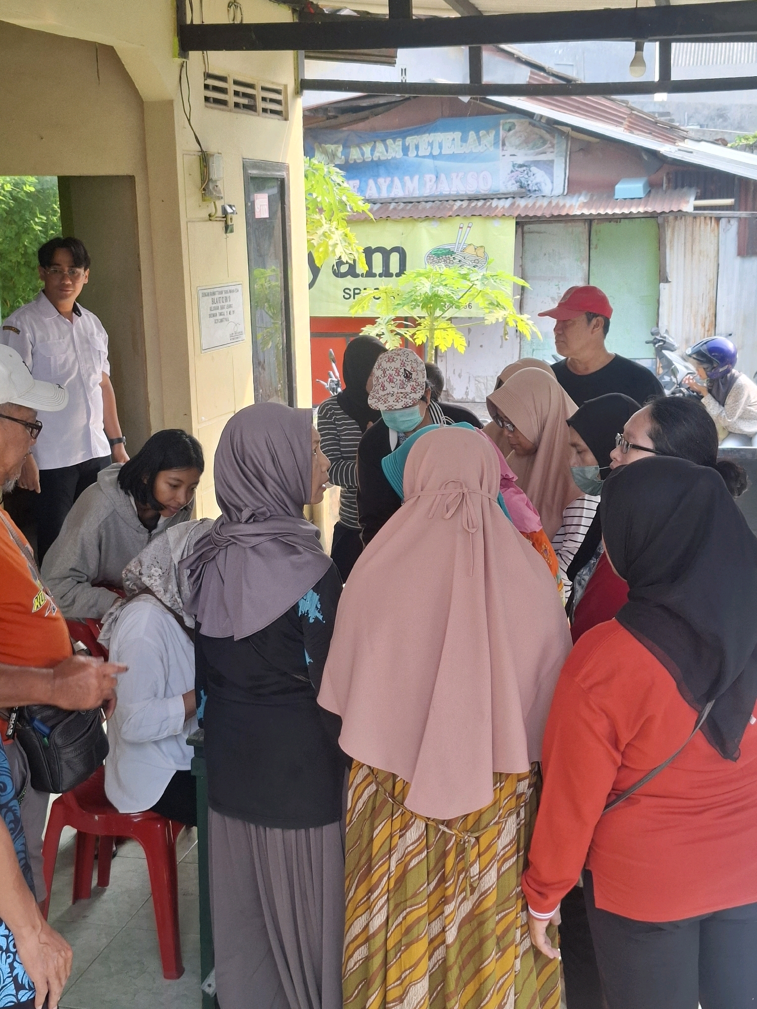 In Surabaya, Indonesia, a group of people are gathered in an outdoor area, possibly a marketplace or a casual eatery. A young man in a white collared shirt and glasses stands to the left, looking towards the camera. In the center, a cluster of individuals, mostly women wearing headscarves, are engaged in conversation. One woman in a grey headscarf is seated on a red chair. Behind them, a man in a red baseball cap observes the scene, and further in the background, a person wearing a blue helmet sits on a motorcycle. The environment appears to be a modest establishment with a corrugated metal roof and a building with louvred windows. Signs with Indonesian text, including "AYAM TETELAN" and "AYAM BAKSO", suggest the presence of food stalls. The overall impression is a candid moment of everyday life in a bustling urban setting.