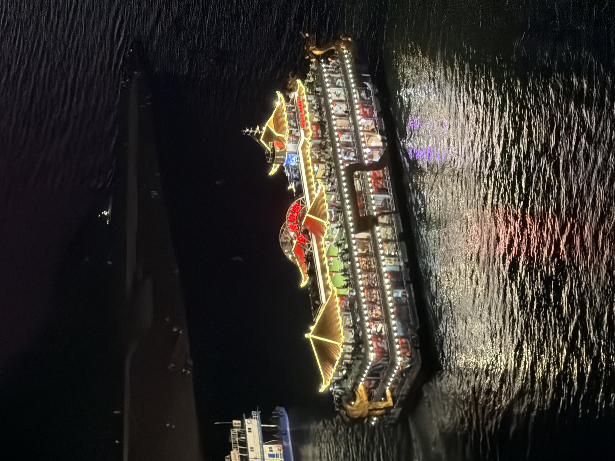 An aerial, eye-level, medium shot captures a brightly illuminated, multi-tiered, traditional Chinese-style junk boat sailing on dark water at night in the Yuzhong District, China. The boat, adorned with golden yellow roofs, red accents, and numerous white and red lights, features ornate dragon carvings on its bow. Multiple levels of windows reveal silhouettes of passengers inside. The water reflects the boat's lights, creating shimmering patterns. In the background, the dark shoreline is visible with faint lights and shadows, suggesting a night scene in a city. Another smaller, white boat is partially visible on the left side of the frame.