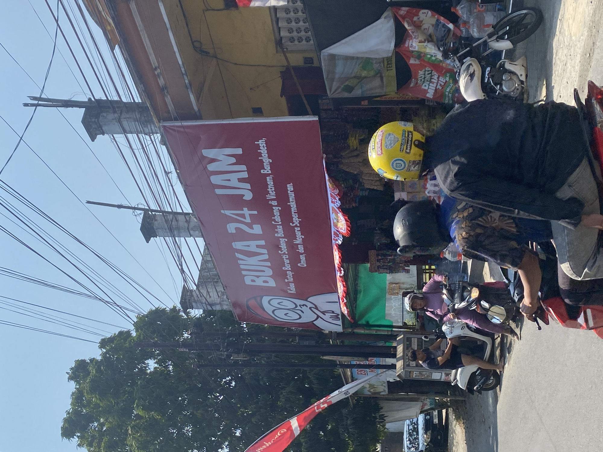 A wide shot captures a street scene in Kediri City, Indonesia, under a clear sky. In the foreground, several people are on motorcycles, facing away from the camera, with one person in a black long-sleeved shirt and grey pants. To the left, a white scooter is parked, with a man in a black t-shirt and shorts standing next to it. A woman in a purple outfit is seated on the scooter, wearing a white helmet. Further back, a white motorcycle is parked on the right side of the image. Above the street, a large red banner reads "BUKA 24 JAM" (Open 24 Hours) in white text, featuring a cartoon character. Below the banner, a storefront with various goods is visible. A red and white flag is displayed on the left. The background shows buildings, trees, and numerous power lines strung across the sky. A dark-colored car is parked on the left side of the road.