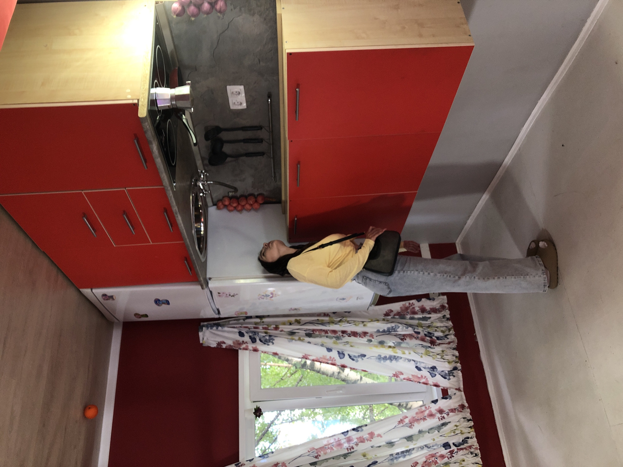 The image shows an upside-down view of a kitchen in Izhevsk, Russia. A young woman is lying on the floor, her body positioned as if she were standing against a wall. She is wearing a yellow long-sleeved shirt and blue jeans. A black purse is slung over her shoulder.
The kitchen features red cabinets and a wooden countertop. A stovetop with two burners is visible. On the wall above the stovetop, a rack holds kitchen utensils, and a pot with a lid is placed on the stove. A sink with a faucet is also visible. To the left of the stovetop, a bunch of red onions hangs. A small orange ball is on the floor to the far left of the image.
In the background, a white refrigerator is adorned with colorful stickers. Floral curtains frame a window that looks out onto a green, leafy environment, suggesting it is daytime and likely spring or summer. The overall impression is that of a playful, surreal environment, possibly an attraction or art installation designed to create an inverted reality.