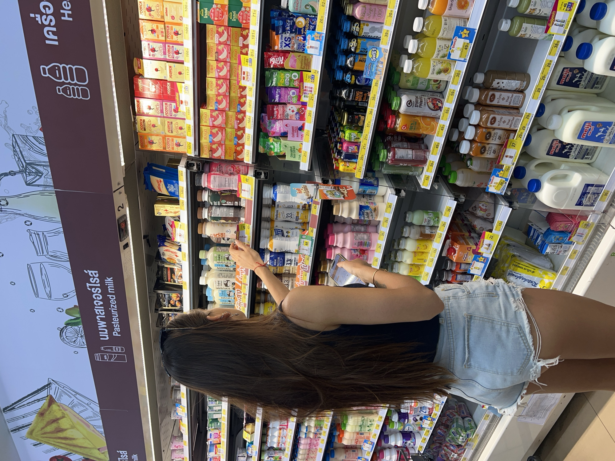 An image depicts a supermarket aisle filled with various packaged beverages and dairy products, viewed from behind a woman. The woman, with long dark hair, wears a dark sleeveless top and light denim shorts. She is standing in front of refrigerated shelves, appearing to browse products with her right hand extended towards an upper shelf, while holding a smartphone in her left hand.
The shelves are brightly lit and neatly stocked with numerous items, predominantly drinks like milk, yogurt drinks, fruit juices, and health supplements. Products are arranged by brand and type, with clear price labels visible below many items.
Notable text includes large signs in Thai and English: "นมพาสเจอร์ไรส์ Pasteurized milk" on a brown vertical sign with milk bottle illustrations, and "เครื่อ He" which likely indicates a healthy drinks section. Specific brands and product types visible through OCR and direct observation include "SCOTCH GOLD", "Jelly me" (with varieties like Collagen, Fiber, Zero, XS G'nite, Chlorophyll Rose Extract), "Herbi", "BRANDS", "Shin S", "meiji", "TOFUSAN", "DAIRYGOLD", "HIGH Protein" drinks, and "Thai Milk Tea". Many labels also show promotional text such as "ราคาพิเศษ" (Special price), "จัดหนัก" (Big deal), "จับคู่ถูกกว่า" (Buy 2, cheaper), and "เติมโปรตีน" (Add protein). The environment suggests a grocery store or convenience store in Thailand.