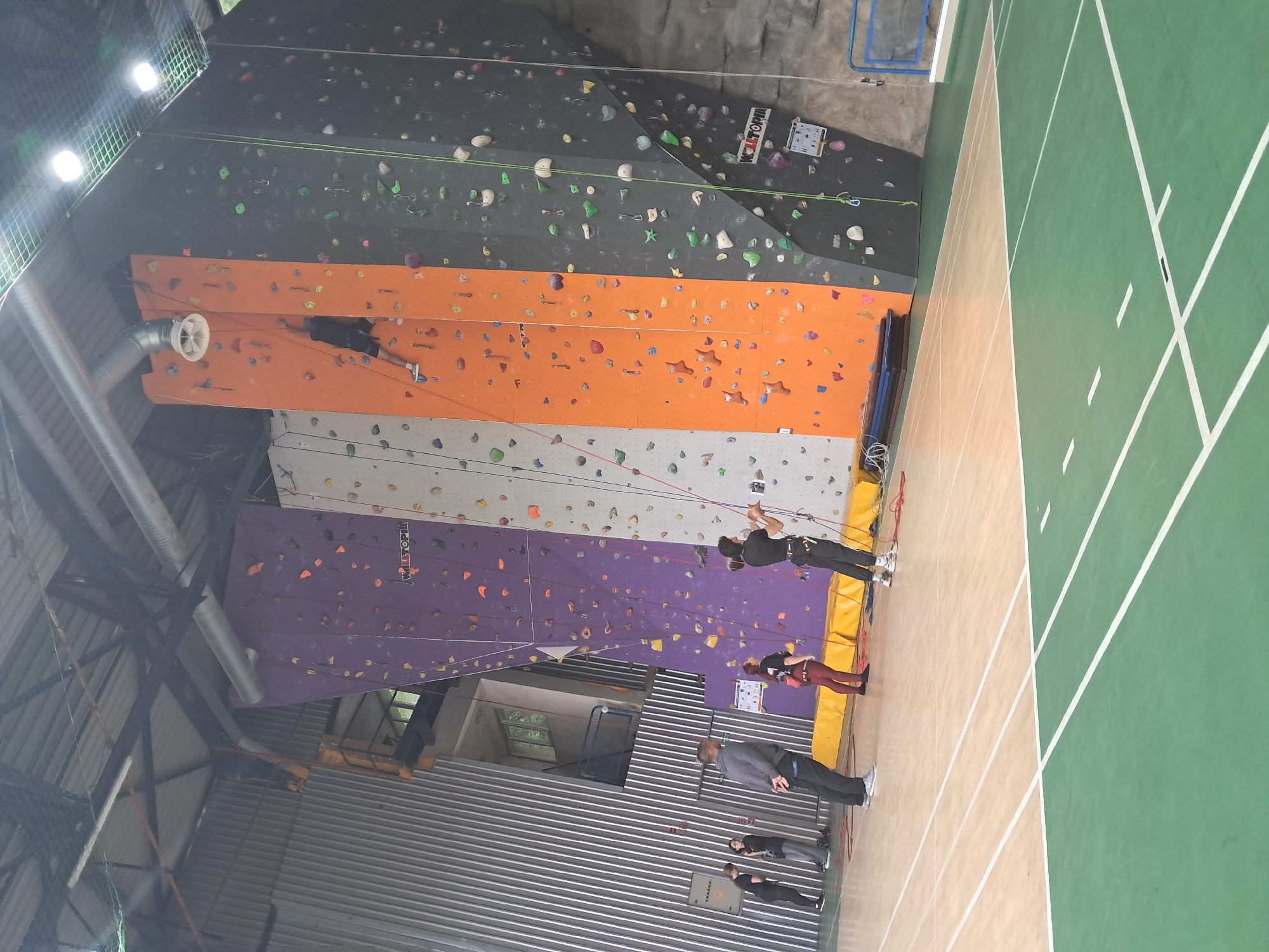 This image captures an indoor sports facility, presenting a multi-colored climbing gym alongside a sports court, viewed from a rotated perspective (originally portrait, rotated clockwise).
The scene is dominated by a series of high climbing walls featuring various sections in black, orange, white, and purple, all covered with numerous brightly colored climbing holds. Climbing ropes are visible, extending up the walls. At the base of the purple wall, yellow crash mats are laid out.
Several people are present, indicating ongoing activity. One individual, wearing a black top and dark shorts/trousers, is actively climbing the orange wall section. At the base of the purple wall, a person in a black top and dark harness is standing, likely belaying or preparing to climb. Nearby, another person in a dark red top and leggings is also standing at the base. Further to the right, a man in a grey jacket and dark trousers observes the scene, while two other individuals in dark clothing stand behind him.
The facility has high ceilings with exposed metal beams and corrugated metal paneling, typical of an industrial-style building. Bright overhead lights, some partially obscured by a protective net, illuminate the area. A large circular ventilation unit is mounted on an upper wall section. The floor to the right consists of a sports court with distinct green and beige/brown sections, marked with white lines.
Visible text includes "VELITOPIA" on white signs attached to both the black and purple climbing walls, identifying the facility. A white information board with numbers and colored squares is also mounted on the black wall. The overall impression is that of a bustling indoor sports center for climbing and other activities.