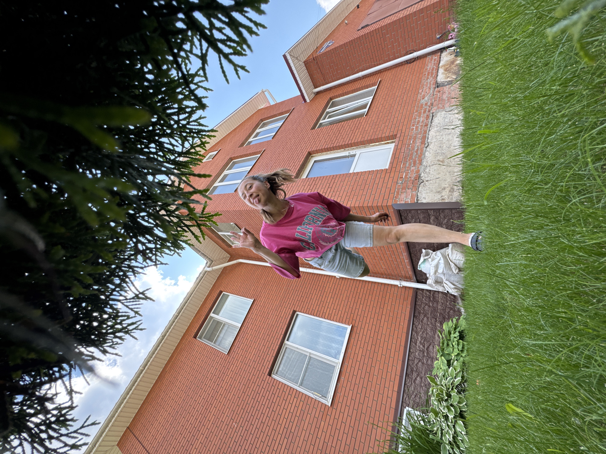 In the city of Perm, Russia, a young woman is pictured in front of a multi-story brick house. She is wearing a pink t-shirt and denim shorts, and she is sticking her tongue out at the camera while holding up two fingers. The house has several windows and a white-trimmed roof. The foreground is filled with lush green grass, and there are some plants and a white sack in the background. The image is taken from a low angle, looking up towards the house and the woman, with tree branches obscuring the top portion of the frame.