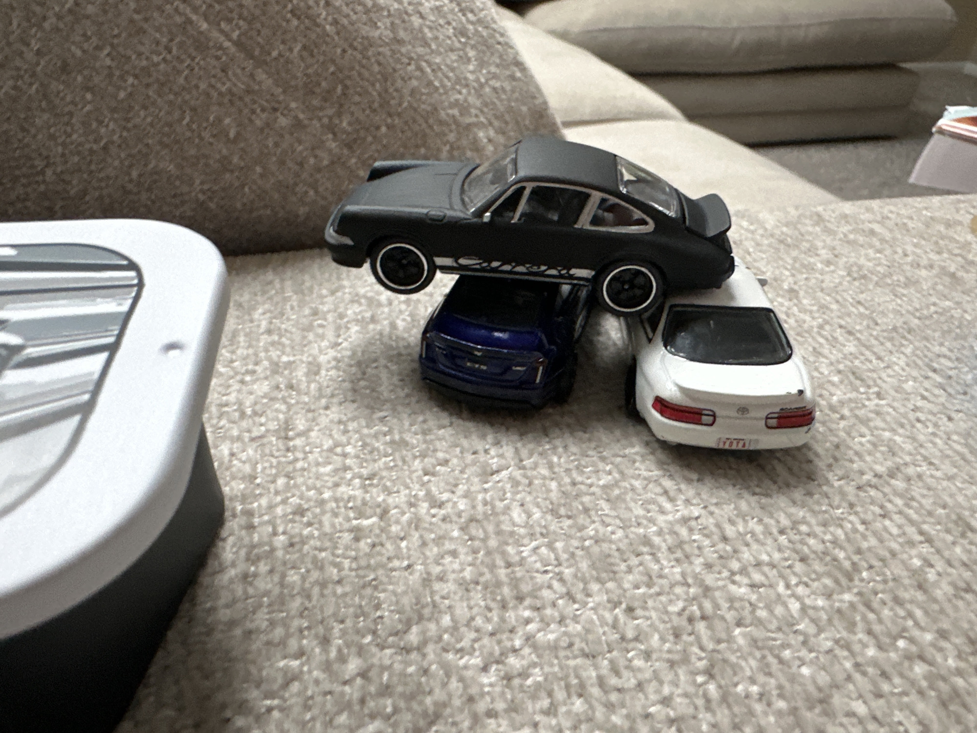 A black toy Porsche 911 Carrera is perched atop a blue toy Cadillac. To the right, a white toy Lexus SC is positioned slightly lower. The toys are resting on a textured, light-colored carpet. In the foreground left, a portion of a white and gray electronic device is visible. The background is out of focus, showing soft furniture. The scene appears to be indoors, possibly in a residential setting.