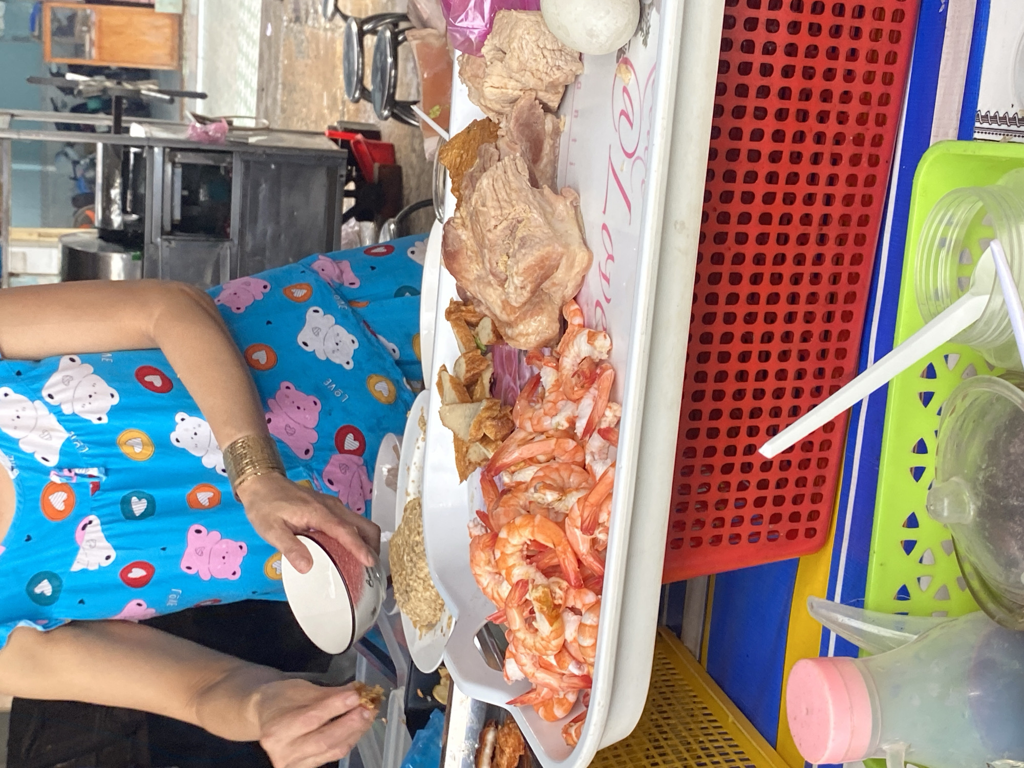 A close-up, eye-level shot shows a person in a blue pajama-like garment with a cartoon bear pattern, preparing food at an outdoor food stall. The person's left hand holds a white bowl with a red rim, while their right hand is picking up some food from a white platter. The platter contains a large mound of cooked shrimp, pieces of fried meat, and what appears to be fried dough. Behind the platter, a white plate holds a pile of cooked oats or noodles. In the background, a stainless steel food cart with a glass display case is visible, along with several stools and the suggestion of a busy street environment. The foreground shows the edge of a red plastic basket, a yellow plastic basket, and a green plastic basket containing bottles and jars of condiments, and a white plastic spoon. The overall scene depicts a street food preparation setting.