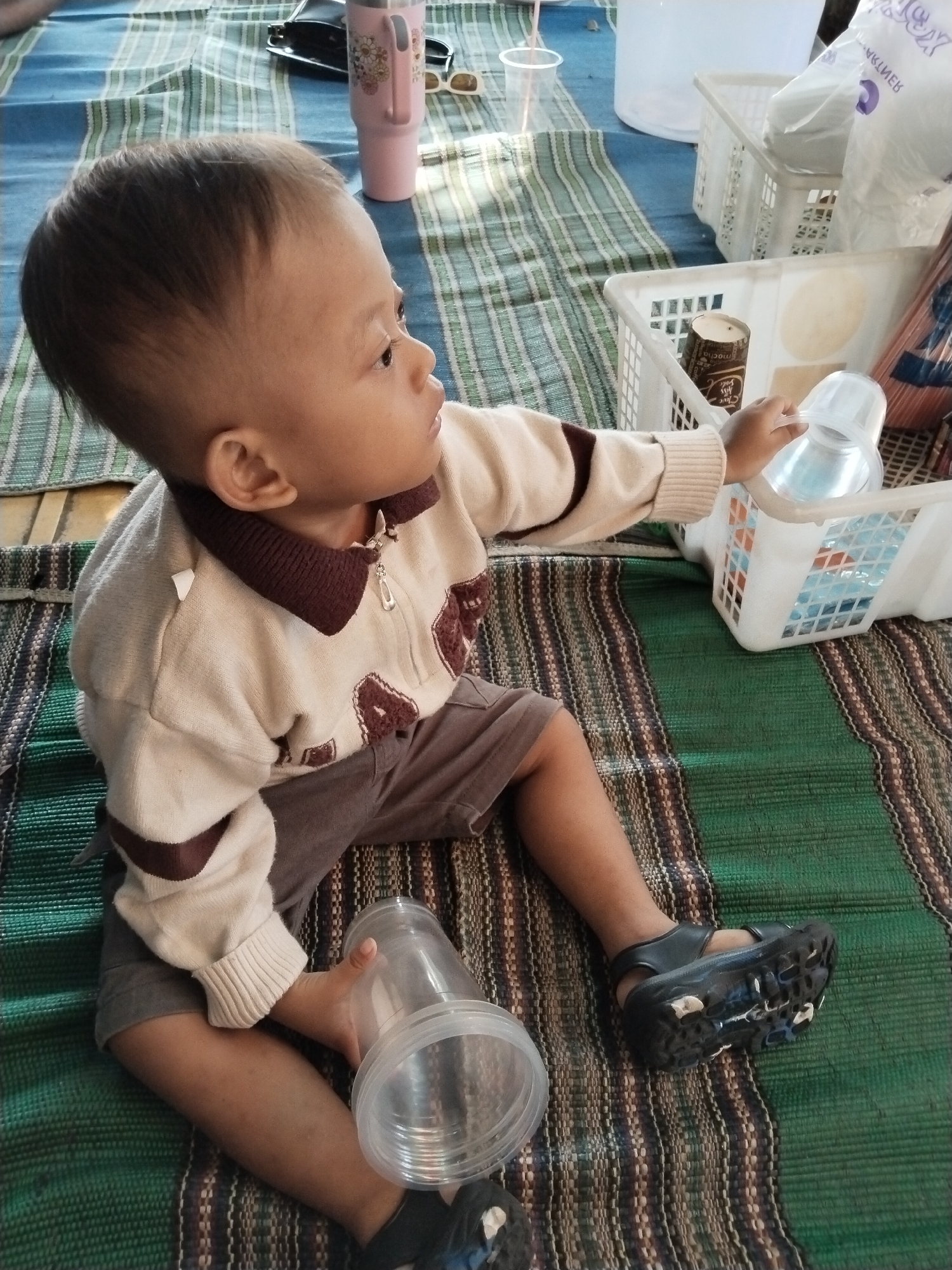 A young child, likely a toddler, is seated on a woven mat with green, blue, and brown stripes. The child is wearing a cream-colored sweater with a dark brown collar and maroon trim, and gray shorts. The child is looking to the right and holding a clear plastic cup with a handle. Nearby, on the mat, is another larger clear plastic cup. In the background, white plastic baskets are visible, one containing what appears to be a disposable coffee cup. A pink thermal mug and a pair of sunglasses are also present on the mat. The scene suggests an outdoor setting, possibly a yard or a public area in Kecamatan Pati, Indonesia, given the local woven mat.