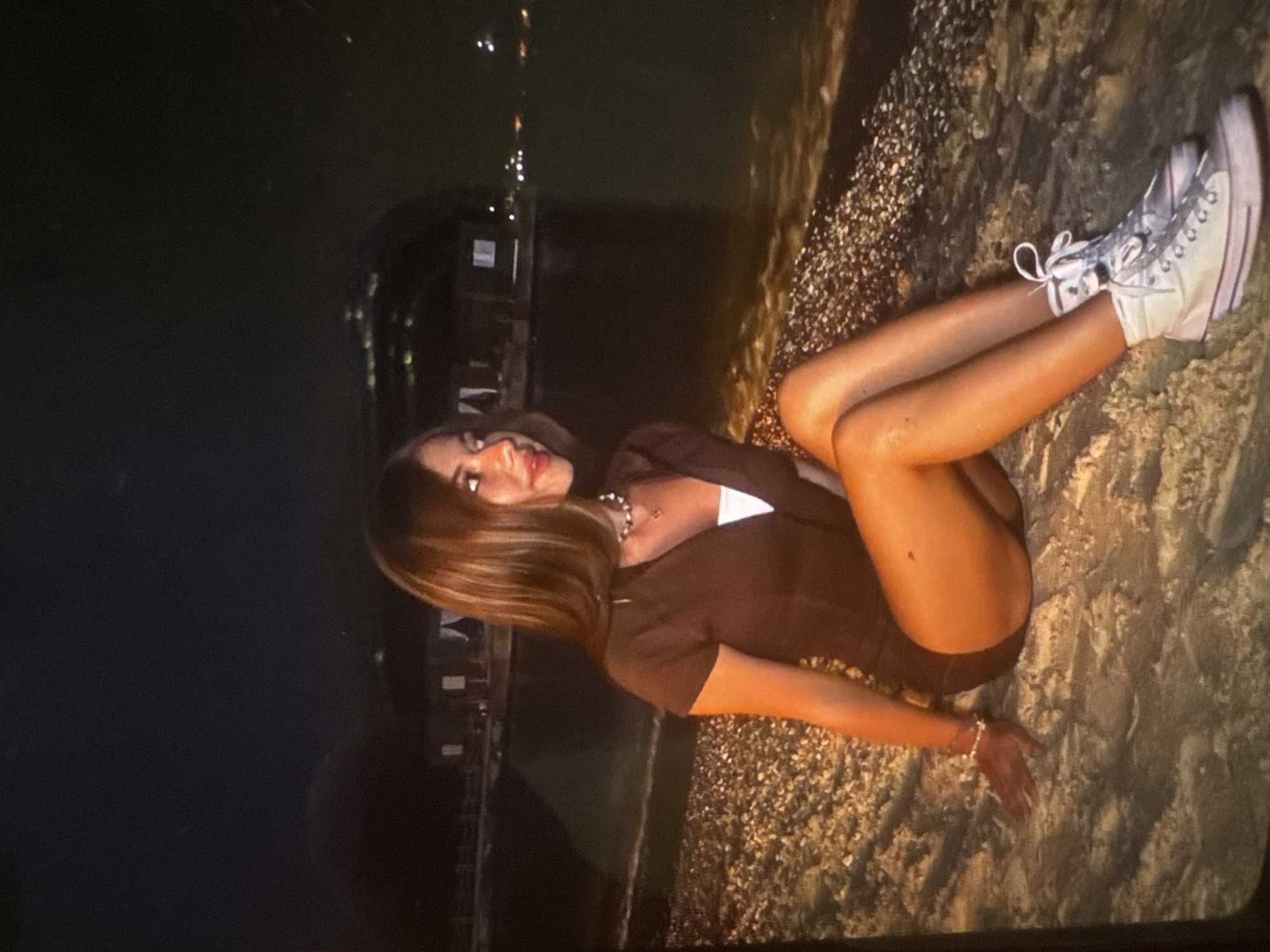 The image displays a young woman posing outdoors at night, lying or reclining on a pebbly and sandy surface, likely a beach or shoreline. A body of dark water is visible behind her, reflecting distant artificial lights from what appears to be a coastal town or structures.
The woman has long, straight brown hair and is looking towards the camera with a slight smile. She is wearing a dark, short-sleeved top or romper that exposes her legs. Her accessories include a light-colored beaded necklace and a bracelet on her right wrist. She has white high-top sneakers with white laces on her feet, with her legs extended.
The time of day is clearly nighttime, as indicated by the dark sky and the array of illuminated buildings and reflections across the water. The setting appears to be a tranquil outdoor environment by the water. No visible text is present in the image.