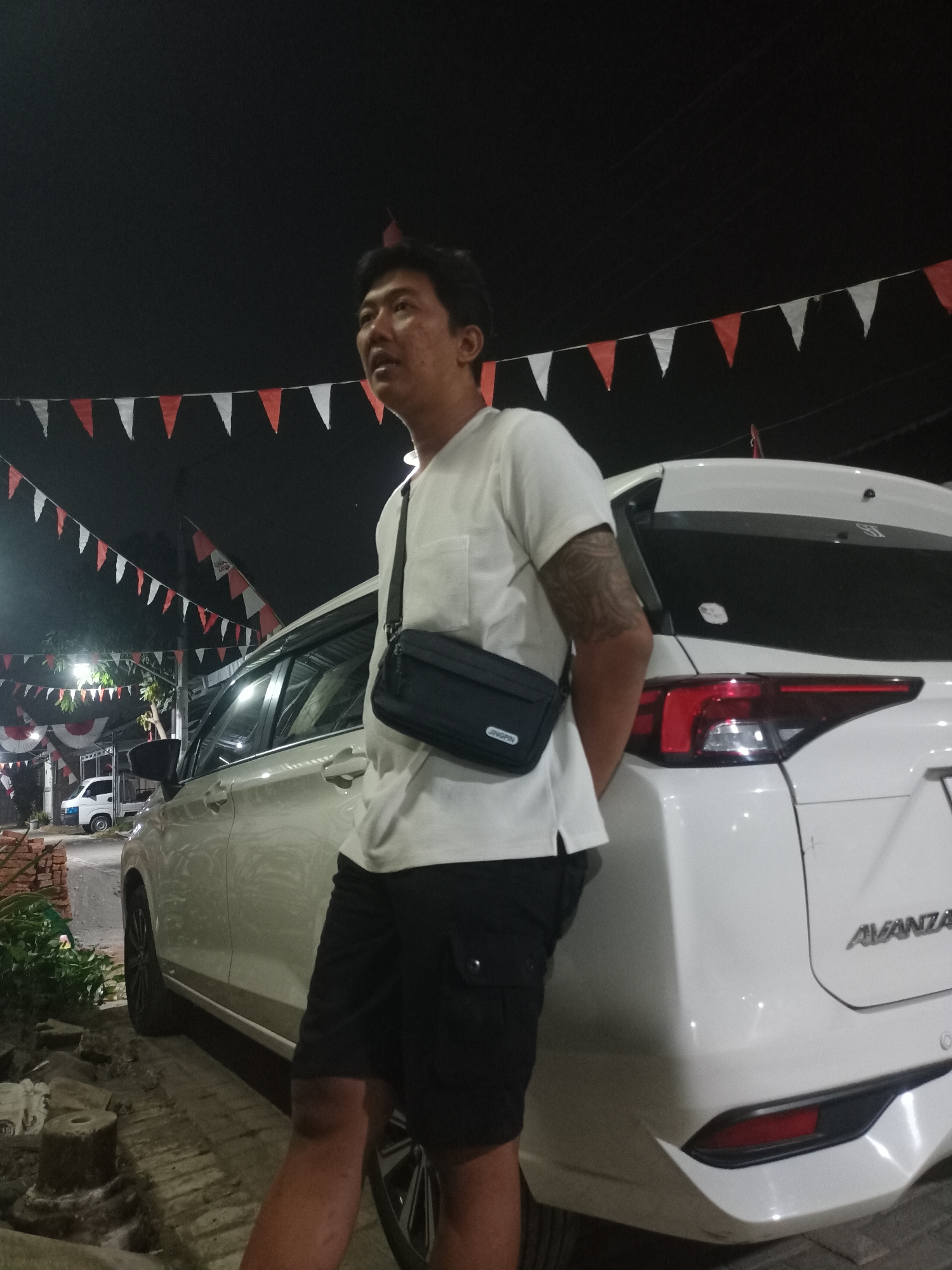 In Mranggen, Indonesia, at night, a man wearing a white t-shirt and black shorts leans against a white Avanza car. He has a black sling bag across his chest and tattoos on his left arm. The car is parked on a brick-paved area, with red and white triangular flags strung across the background. Trees and lights are visible in the distance, and a white truck is parked further down the street. The scene appears to be outdoors, possibly a residential area or a small business.