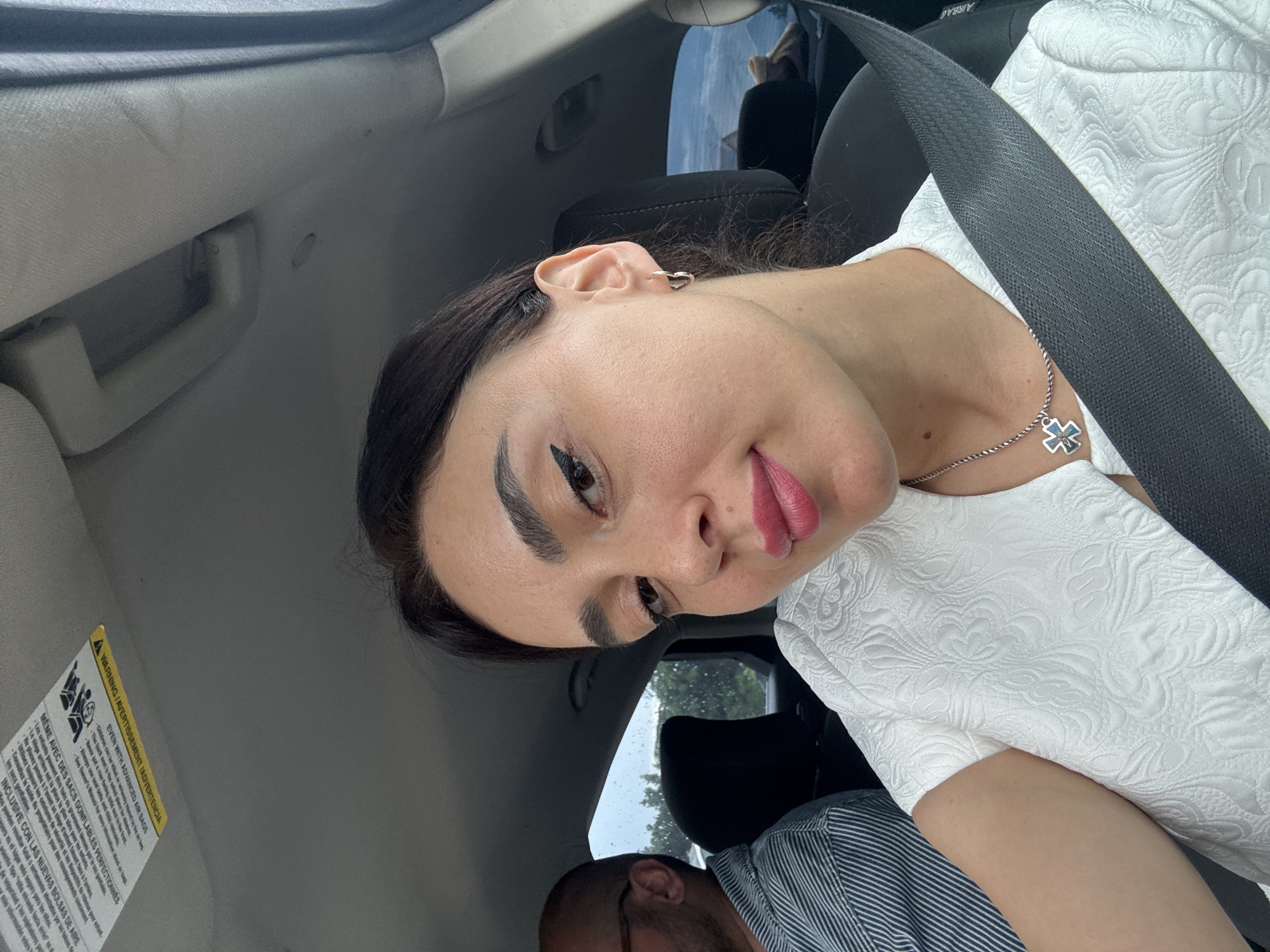 A woman and a man are in a car. The woman is in the passenger seat, and the man is in the driver's seat. The woman is looking at the camera and smiling. She is wearing a white shirt with a textured pattern and a silver necklace with a cross pendant. The man is wearing a striped shirt and glasses. The car appears to be moving, as indicated by the motion blur in the background. The weather outside is overcast, with visible rain on the window. The scene is likely captured in Tbilisi, Georgia, given the context provided.