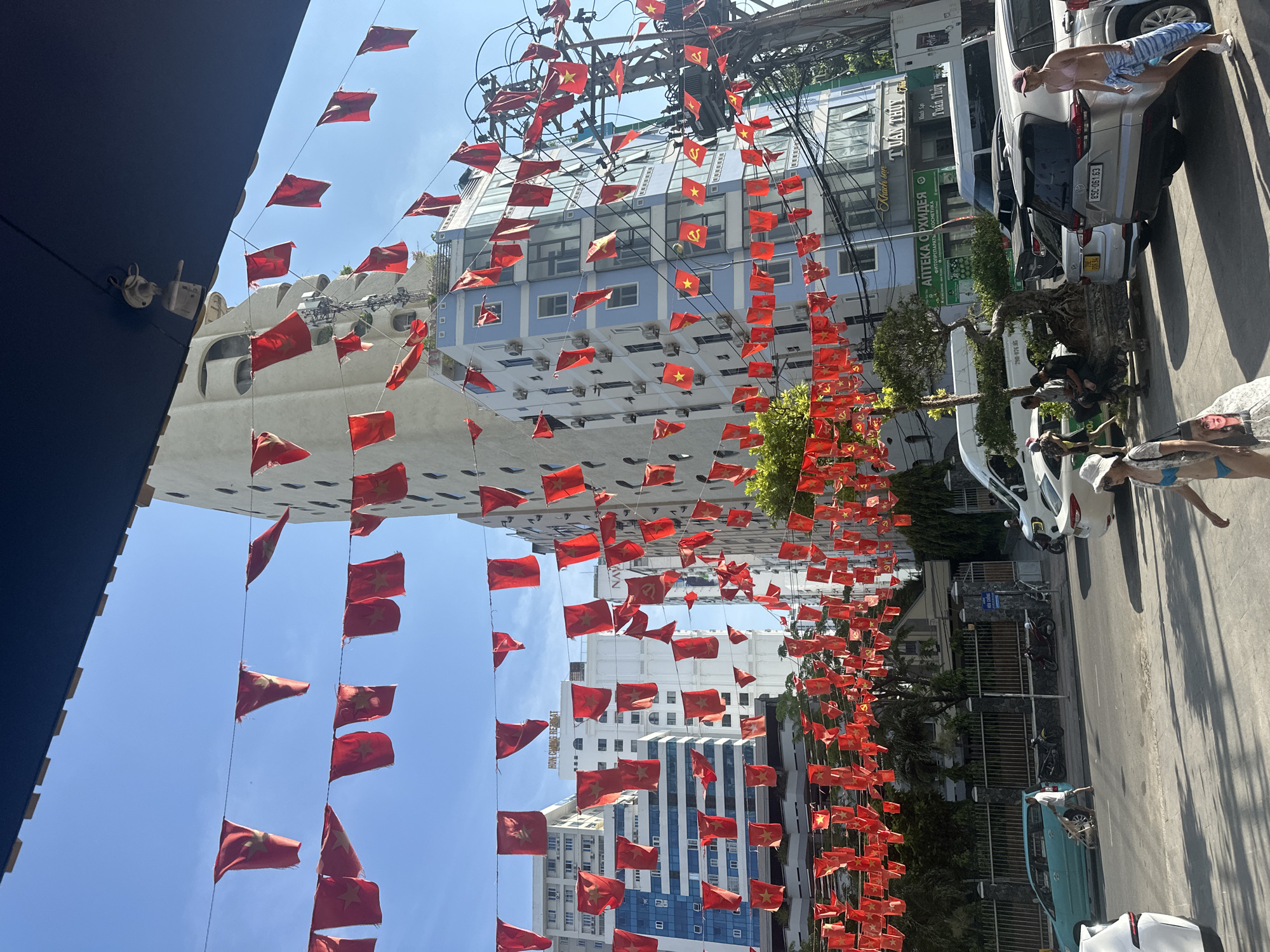The image depicts a street scene in Nha Trang Ward, Vietnam, with numerous red flags bearing a yellow star strung across wires overhead. The flags are a prominent visual element, suggesting a festive or patriotic occasion.
In the background, several multi-story buildings line the street. One tall, light-colored building with an unusual curved architectural style stands out. Adjacent to it is a blue and white building with many windows. Utility poles with a dense network of wires are visible, also adorned with some of the red flags.
The street level shows parked vehicles, including cars and motorcycles. A light blue minivan is parked on the left side of the road, and a silver SUV is prominent in the foreground on the right. A white sedan is also visible.
Several people are present. In the foreground on the right, a woman in a bikini and a sun hat is walking past the silver SUV. Further left, a man in a white t-shirt and shorts is walking a dog. Another man is seated on a bench next to a decorative bonsai tree. Other figures appear in the background, including a person near a white van.
The environment appears to be a sunny day, with clear blue skies. The overall impression is one of an urban street with decorations and daily life unfolding. Signs on the buildings are in Vietnamese, with some Cyrillic script also visible, indicating a mixed cultural influence or a specific business name.