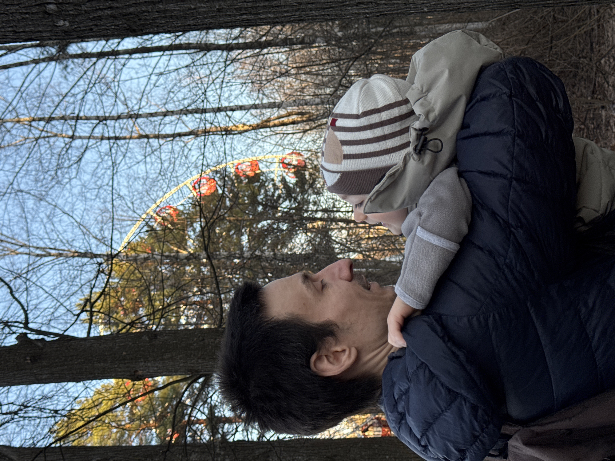 The image, set in Izhevsk, Russia, captures a heartwarming outdoor scene featuring an adult man and a young child. The man, seen in profile on the left, has dark hair and a light mustache. He looks upwards with a joyful or engaged expression, his mouth slightly open as if smiling or talking. He is dressed in a dark blue puffer jacket. On the right, a young child is positioned, likely on the man's shoulders or being held up, looking down towards him. The child wears a light-colored beanie with brown stripes and a distinct brown design, a light-colored hooded jacket, and grey sleeves or gloves.
The background is dominated by a clear, bright blue sky and numerous bare tree branches, indicative of a late autumn or winter season. Through these branches, a large Ferris wheel with bright red gondolas and yellow or gold lights outlining its structure is visible, suggesting the location is an amusement park or fairground. Thick tree trunks are also present on the left side of the frame. The heavy winter clothing worn by both individuals further confirms the cold weather. The interaction between the man and child conveys a sense of warmth, affection, and shared joy within the serene, cold-weather park environment. No visible text is present in the image.