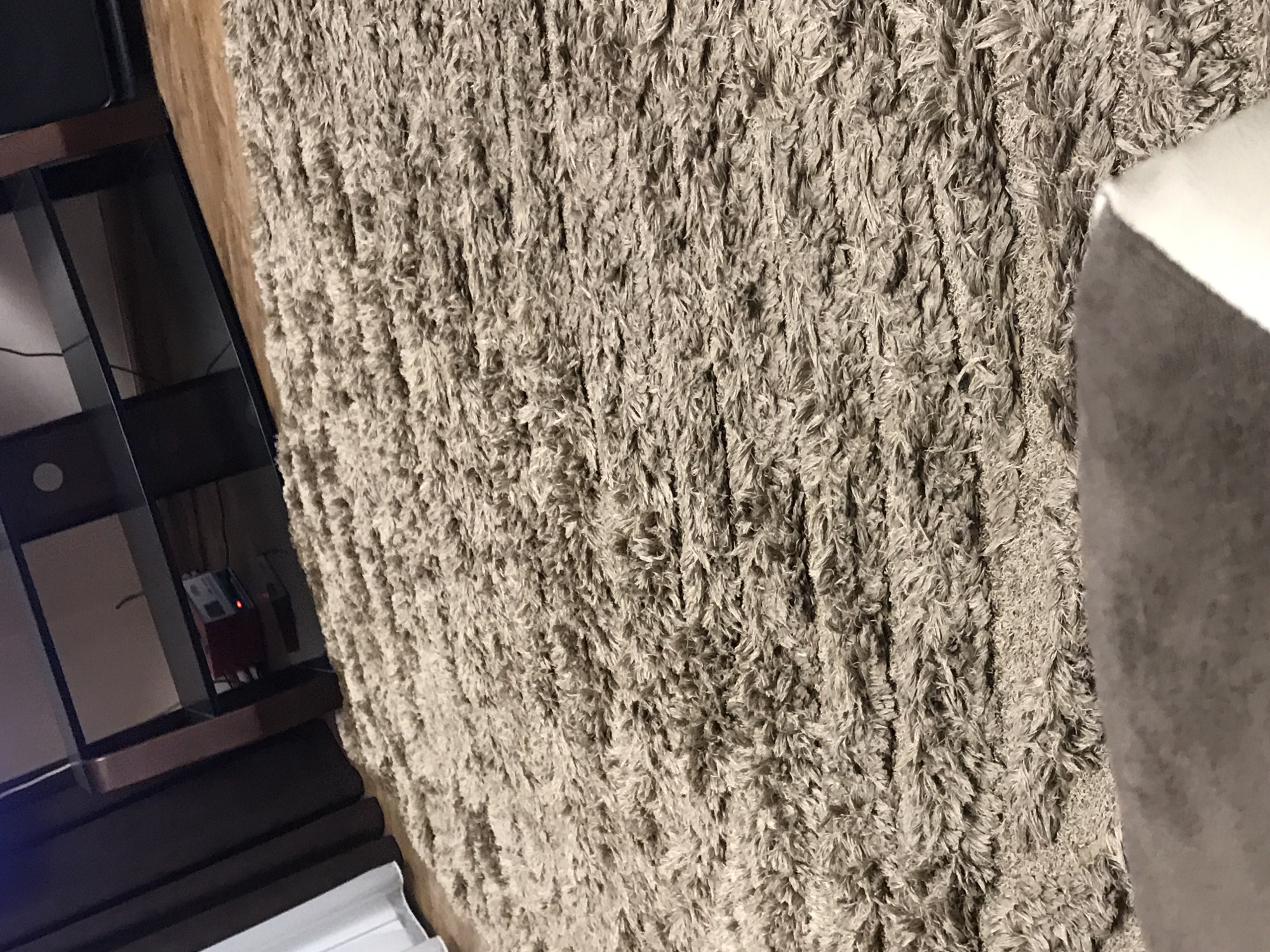 The image shows a close-up of a high-pile carpet in a neutral beige color. To the left of the carpet is a dark-colored shelving unit, possibly for entertainment or display. Several wires and electronic devices are visible within the shelving unit. The right portion of the image shows a portion of a white or light-colored object, possibly a piece of furniture like a sofa or chair. The specific location in Yerevan, Armenia, cannot be determined from the image content.