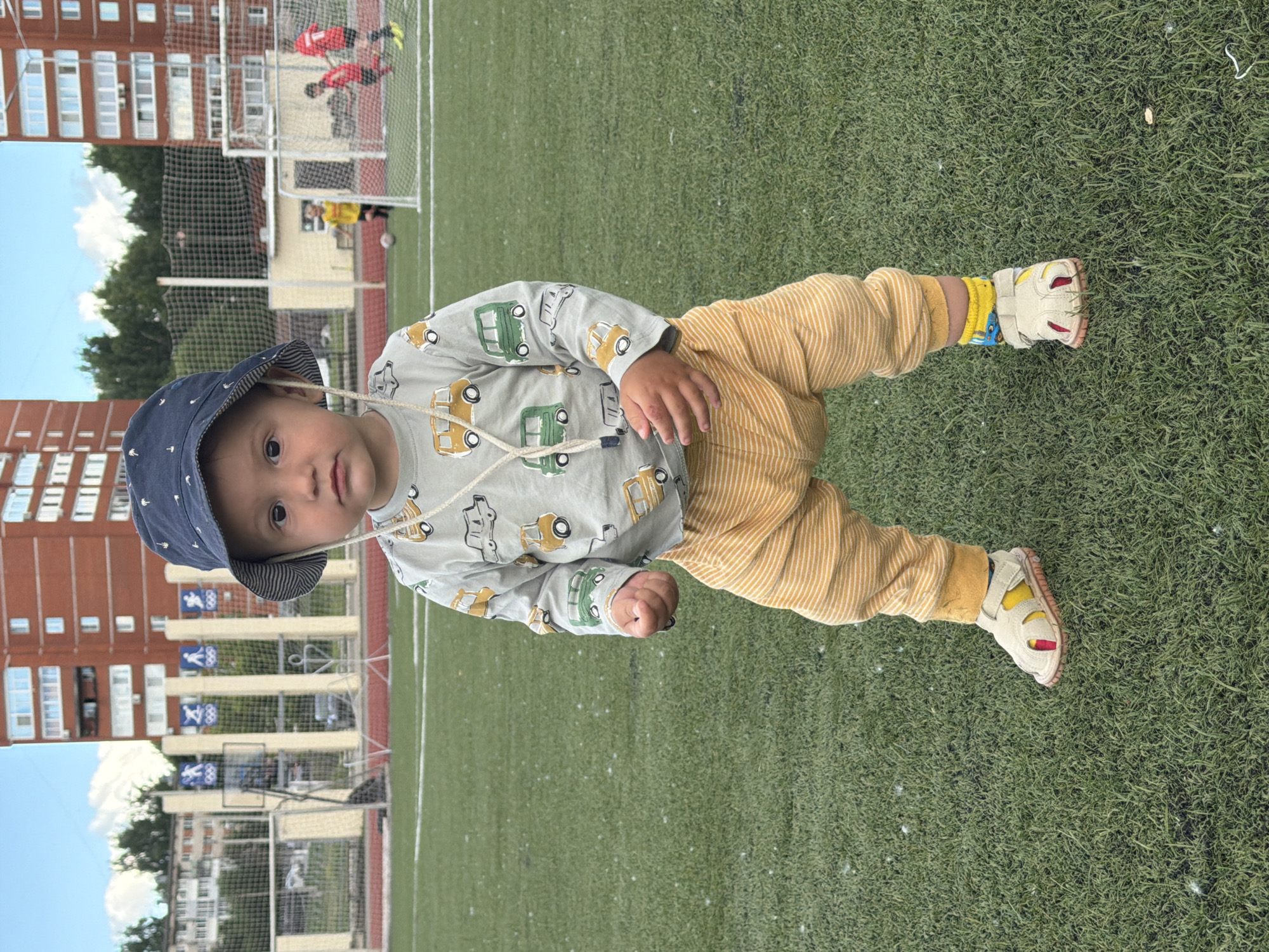 A young child, wearing a light blue bucket hat and a long-sleeved grey shirt with a pattern of yellow and green cars, sits on a vibrant green artificial turf field. The child is also wearing yellow striped pants and white sandals with yellow and red accents, and blue and yellow patterned socks. The child's left hand is clenched into a fist.
In the background, there are tall apartment buildings constructed with red brick. A soccer goal with a net is visible, and two individuals in red jerseys are near it, suggesting a soccer game or practice. A person in a yellow shirt is also present on the field. Several blue banners with white icons, possibly Olympic rings, are displayed on a fence. The sky is a clear blue with scattered white clouds. The lighting suggests it is daytime, likely sunny. The scene appears to be a recreational or sports facility within a residential area. No visible text is discernible.