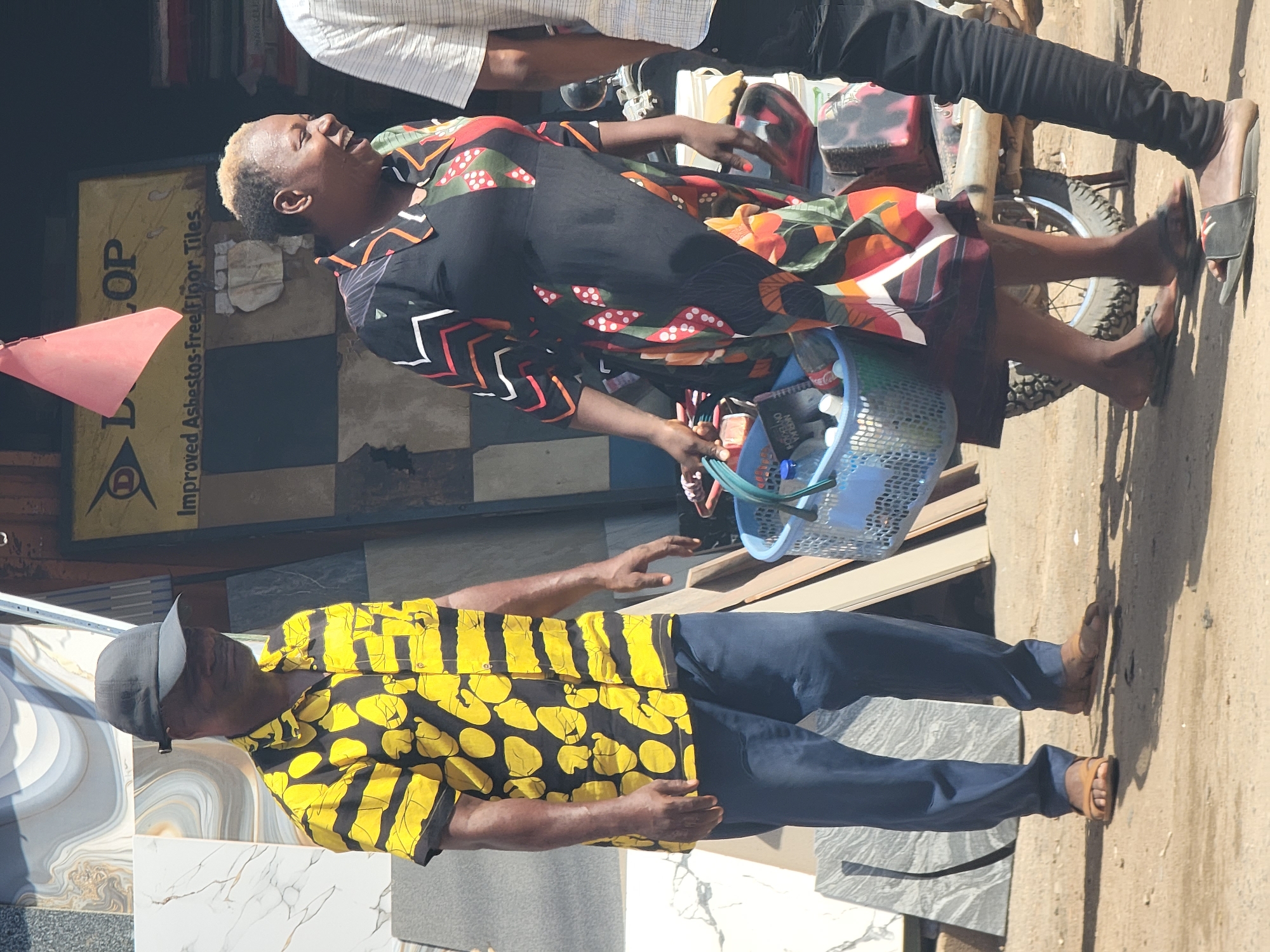 The image captures a street scene in Maiburiji, Nigeria. In the foreground, a man wearing a yellow and black patterned shirt, dark blue pants, and a baseball cap stands on a variety of ceramic tiles. His right arm is outstretched. Behind him, a woman sits on a motorcycle holding a blue plastic basket filled with assorted items. She wears a black shirt with red, orange and green patterns, and a multicolored skirt. Another person in a light-colored shirt and black pants stands beside her. Behind the woman is a wall advertisement for "Improved Asbestos-free Floor Tiles." The ground appears to be dirt and concrete. The photo is rotated clockwise.