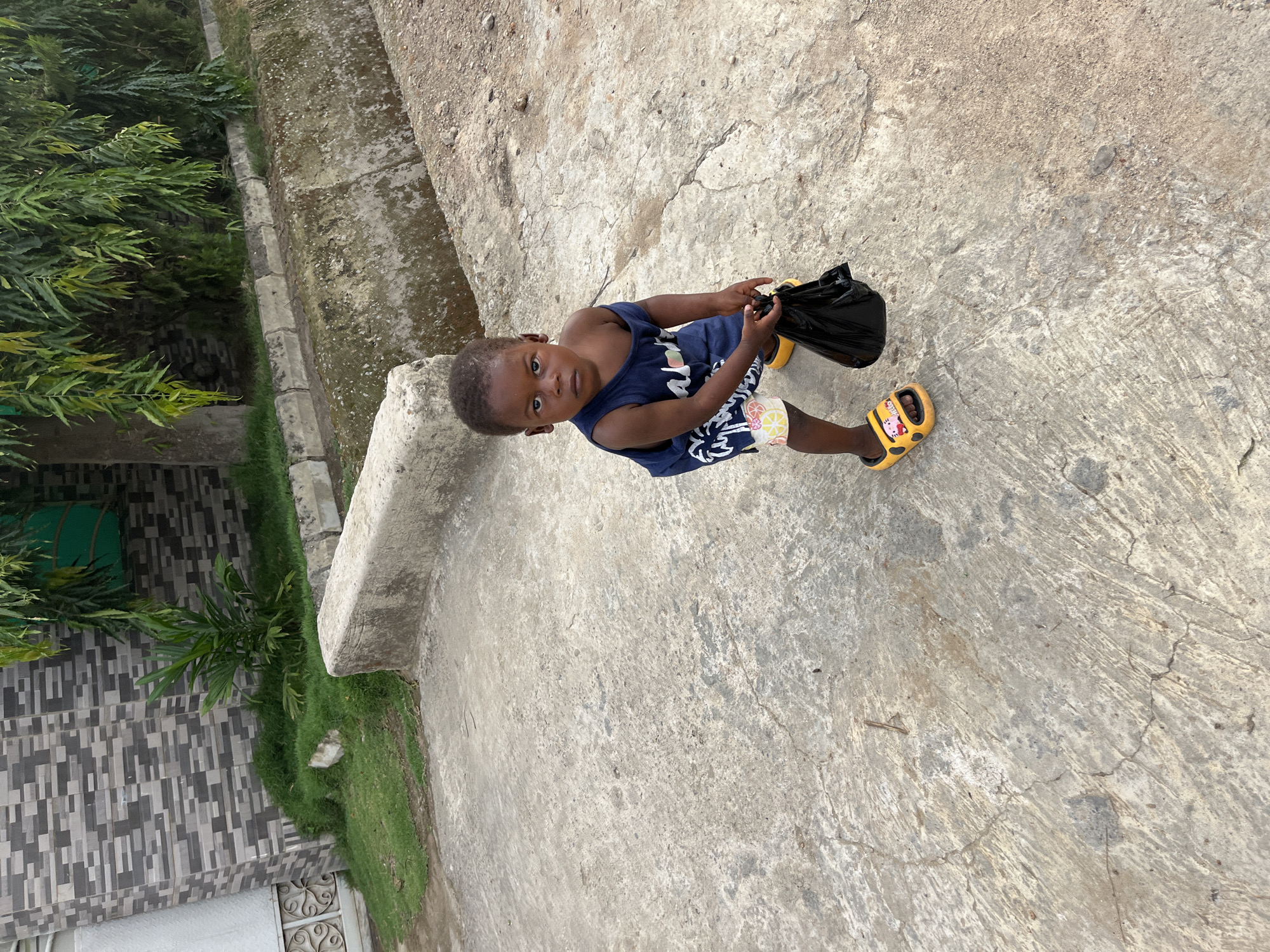 A small child stands on a concrete surface, likely a walkway or patio. The child, who appears to be of African descent, is wearing a blue tank top, patterned shorts, and yellow sandals with a cartoon design. The child is holding a black plastic bag. The background includes green vegetation and parts of a building with light and dark gray tiles. The location is presumed to be Abuja, Nigeria.