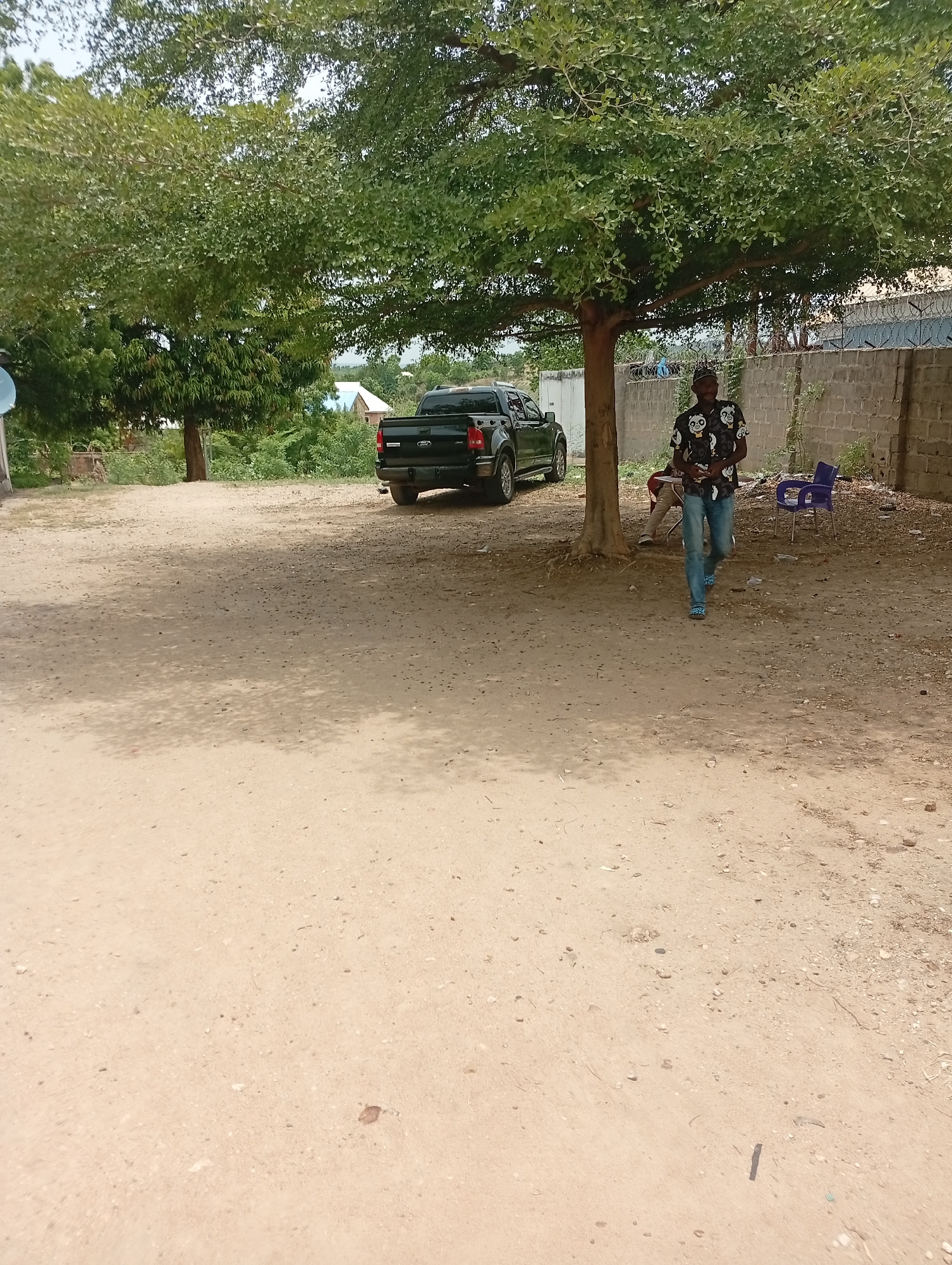 The scene is an open dirt lot in Adankolo, Nigeria, on a bright day. A dark-colored pickup truck is parked under the shade of a large, leafy tree. A man wearing a patterned shirt and jeans walks towards the viewer, away from the tree. Behind the tree are glimpses of a person seated, and a blue plastic chair. A concrete block wall lines part of the lot's perimeter. Another tree is visible in the background, and some buildings in the far distance.