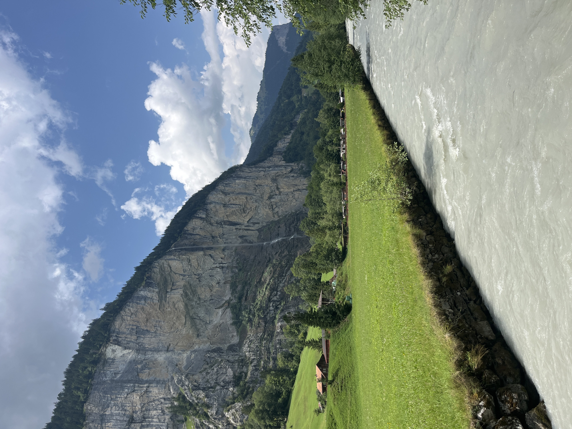A wide, choppy, milky-white river flows from the lower right to the lower left of the frame, bordered by a stone wall topped with a strip of grass. To the left of the river, a vibrant green meadow extends towards a steep, towering cliff face. The cliff is a mix of grey rock and patches of green vegetation, with a thin waterfall cascading down its side. At the base of the cliff, nestled on a grassy incline, are two traditional Swiss chalets with red roofs. Beyond the chalets, a line of trees stretches across the middle ground, with more trees and rolling green hills visible in the background, leading up to a distant, hazy mountain range. The sky above is a bright blue, dotted with fluffy white clouds. This scene depicts the Lauterbrunnen Valley in Wengen, Switzerland, famous for its dramatic cliffs and waterfalls.