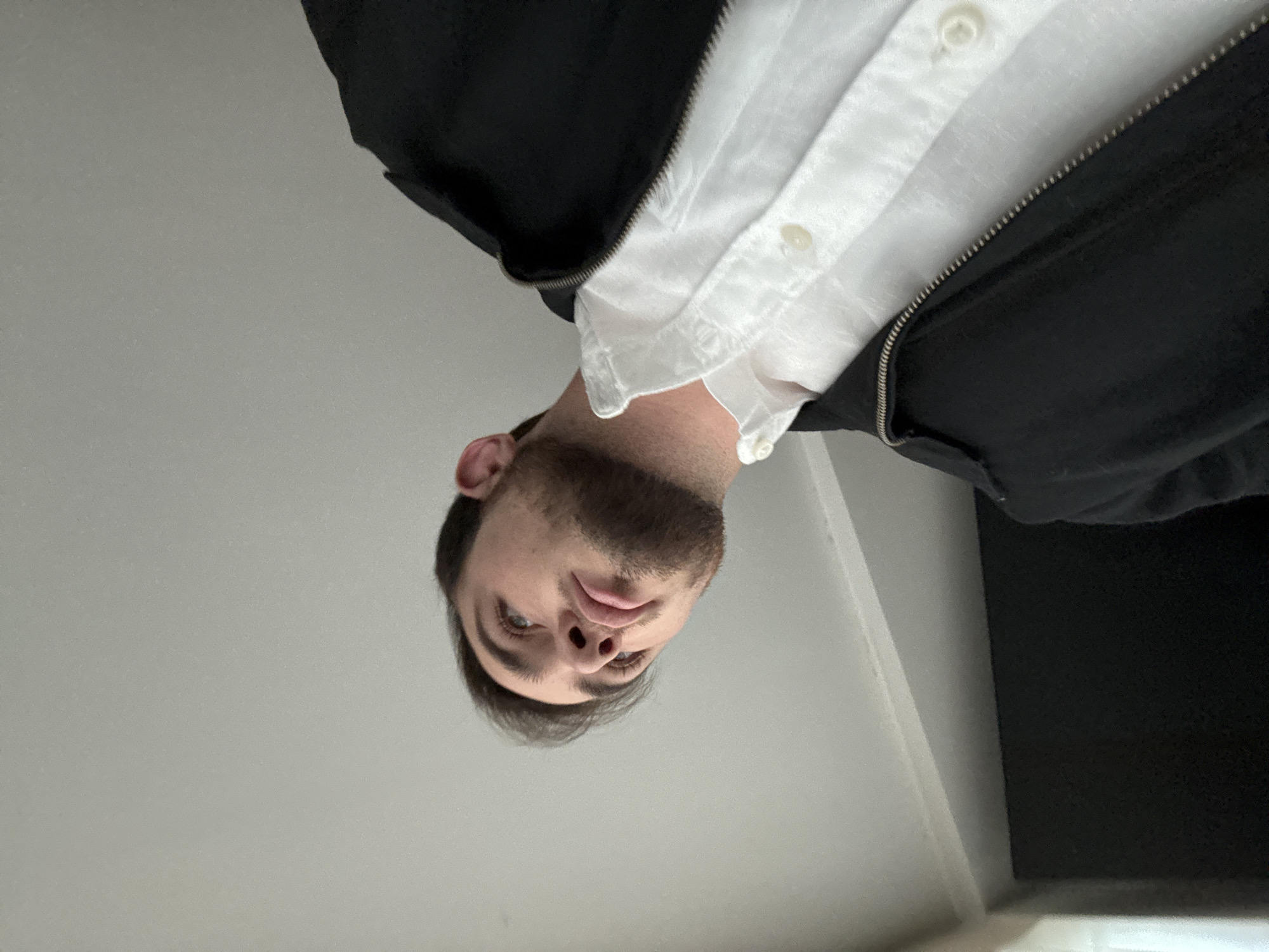 A close-up, low-angle portrait shot captures a young to middle-aged man looking directly downwards into the camera. His head is inverted in the frame, with his face positioned towards the bottom. The man has short, dark hair, a neatly trimmed beard, and striking light-colored eyes. His expression is neutral and direct.
He is wearing a white collared shirt, likely of a textured fabric, underneath an open, dark (possibly black) zippered jacket. The metallic zipper and buttons on the white shirt are visible.
The background is a plain, light grey or off-white surface, indicating an indoor setting, likely a ceiling or an upper wall. A subtle corner is visible towards the top right of the frame. The lighting is soft and even, suggesting ambient indoor light. No specific location cues are visible in the background for San Bernardino Tlaxcalancingo, Mexico, beyond the interior nature of the shot. No text is visible in the image. The photo emphasizes the man's face and upper torso from this unusual perspective.