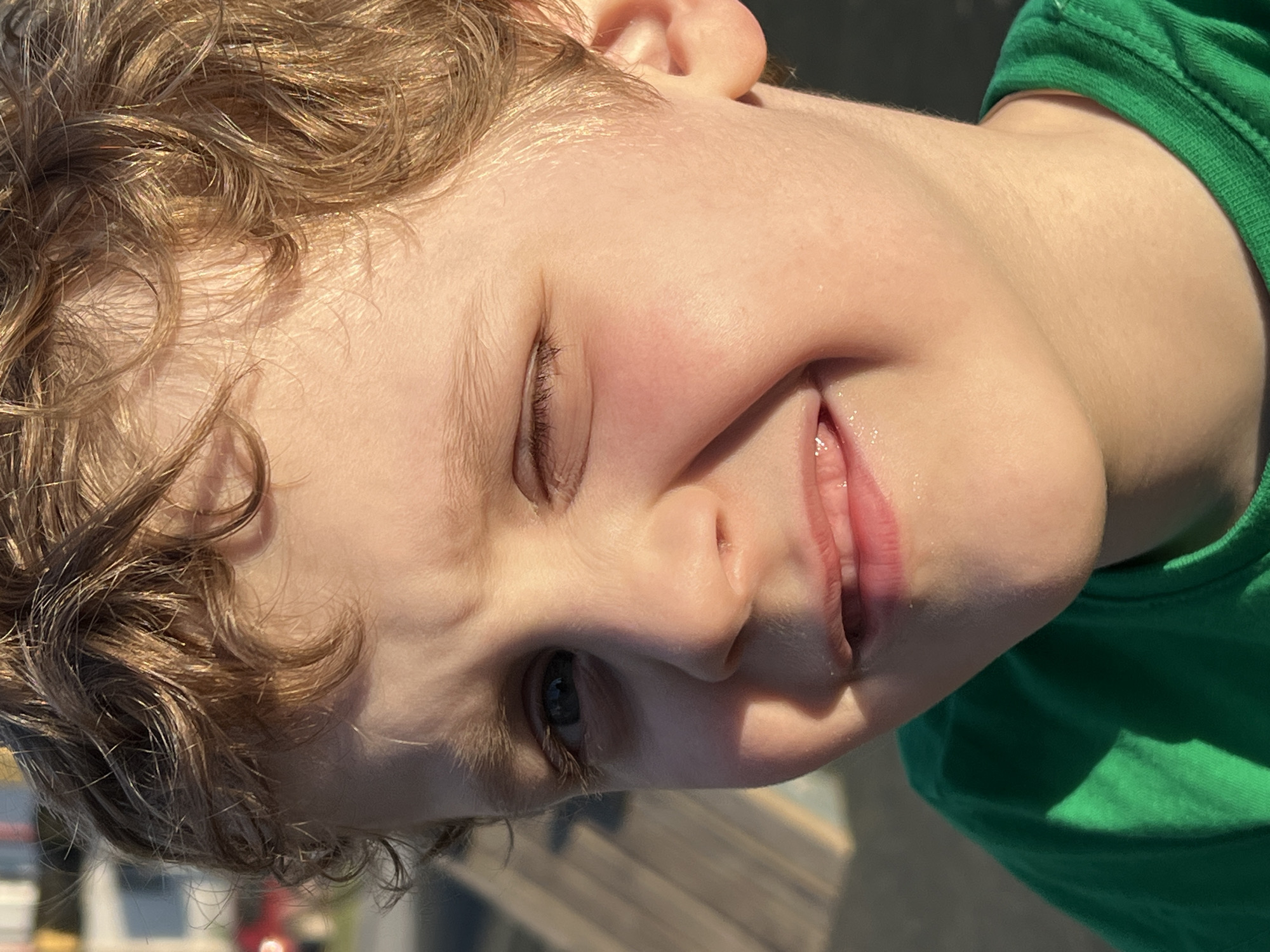 A close-up of a child's face. The child has light skin, curly blonde hair, and one eye open, while the other is closed in a slight squint or wink. The child is smiling slightly, showing a glimpse of their upper teeth. They are wearing a bright green shirt. The background is out of focus, but suggests an outdoor setting, possibly with wooden structures and some indistinct urban elements. There is no definitive evidence of the location being in Noviy, Russia.