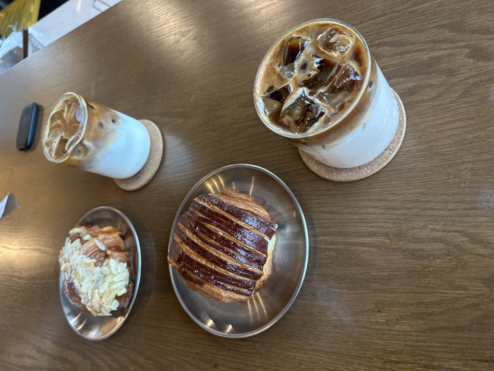 A high-angle, medium-shot photograph shows a wooden table with two glasses of iced coffee, two pastries, and a black object. The coffee drinks are in cylindrical glasses with ice, coffee, and a white liquid (likely milk). Both are resting on small, round, cork coasters. The pastry closest to the lower left is an almond croissant, while the other is a plain croissant with glazed, striped dark brown topping. Both pastries are displayed on small, oval, metal dishes. To the left of the lower coffee drink is a black, rectangular object. The scene is possibly set in a cafe or restaurant in San Pedro Garza García, Mexico. No people are visible in the image.