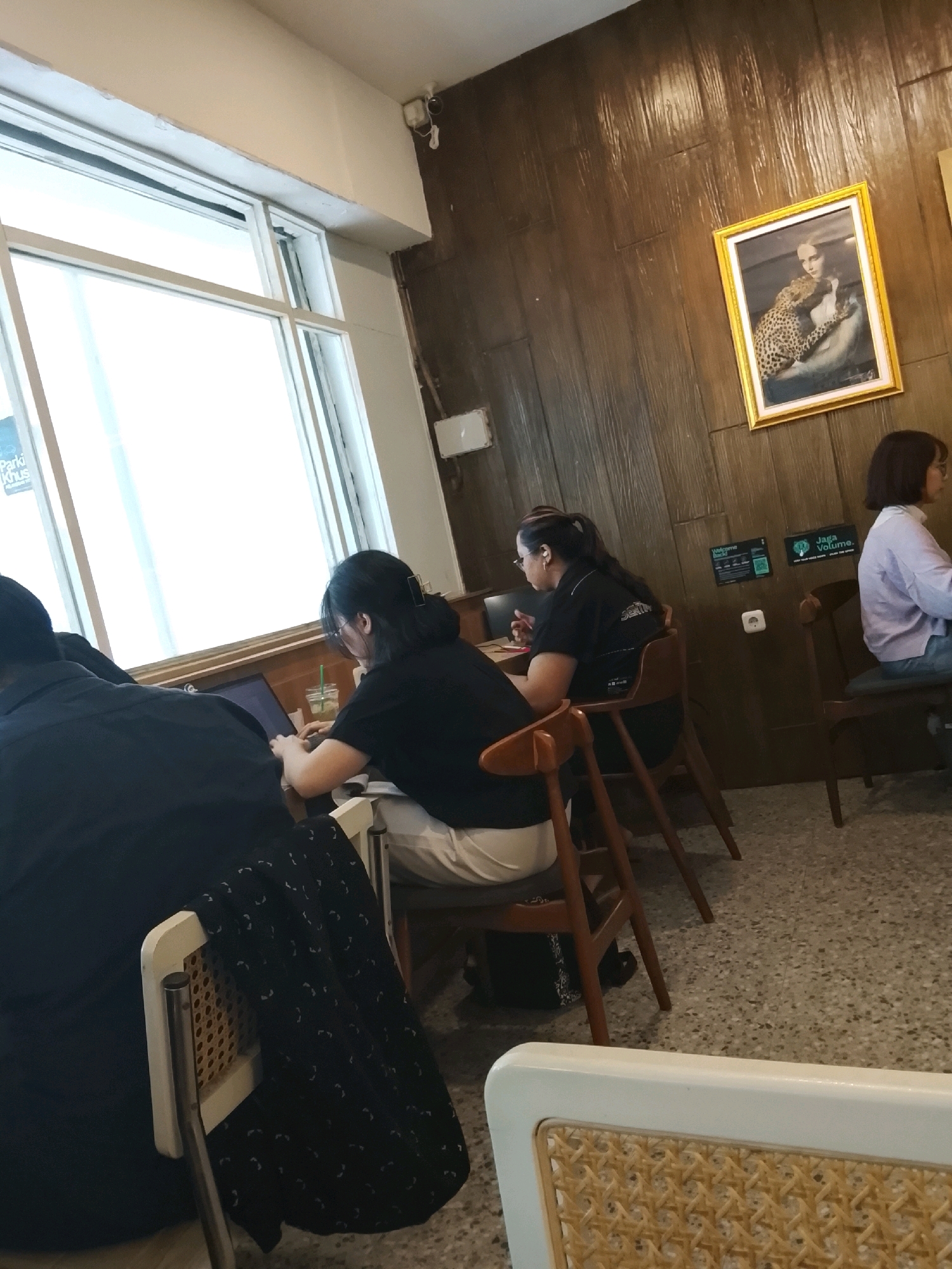 The image depicts an indoor scene, likely a cafe, with several individuals present. In the foreground, a person wearing a dark blue shirt is seated at a table, their back to the camera. To their right, a woman with dark hair is working on a laptop, also seated at a table. She is wearing a black shirt and white pants and has a drink with a straw in front of her. Further to the right, another woman with dark hair and glasses is seated at a table, wearing a black polo shirt and white pants. She is looking at something on her lap. In the background, a woman with short dark hair and glasses is seated at a table, wearing a light purple long-sleeved shirt and blue jeans. She is looking to her right.
The environment features wooden-paneled walls and a large window on the left side. A framed picture of a woman with a leopard hangs on the wall. There are tables and chairs throughout the space. The floor is a speckled grey tile. Signs are visible on the wall, including one that says "Welcome Back!" and another with "Jaga Volume." A security camera is mounted on the ceiling near the window. Based on the provided information, this scene is located in Surabaya, Indonesia.