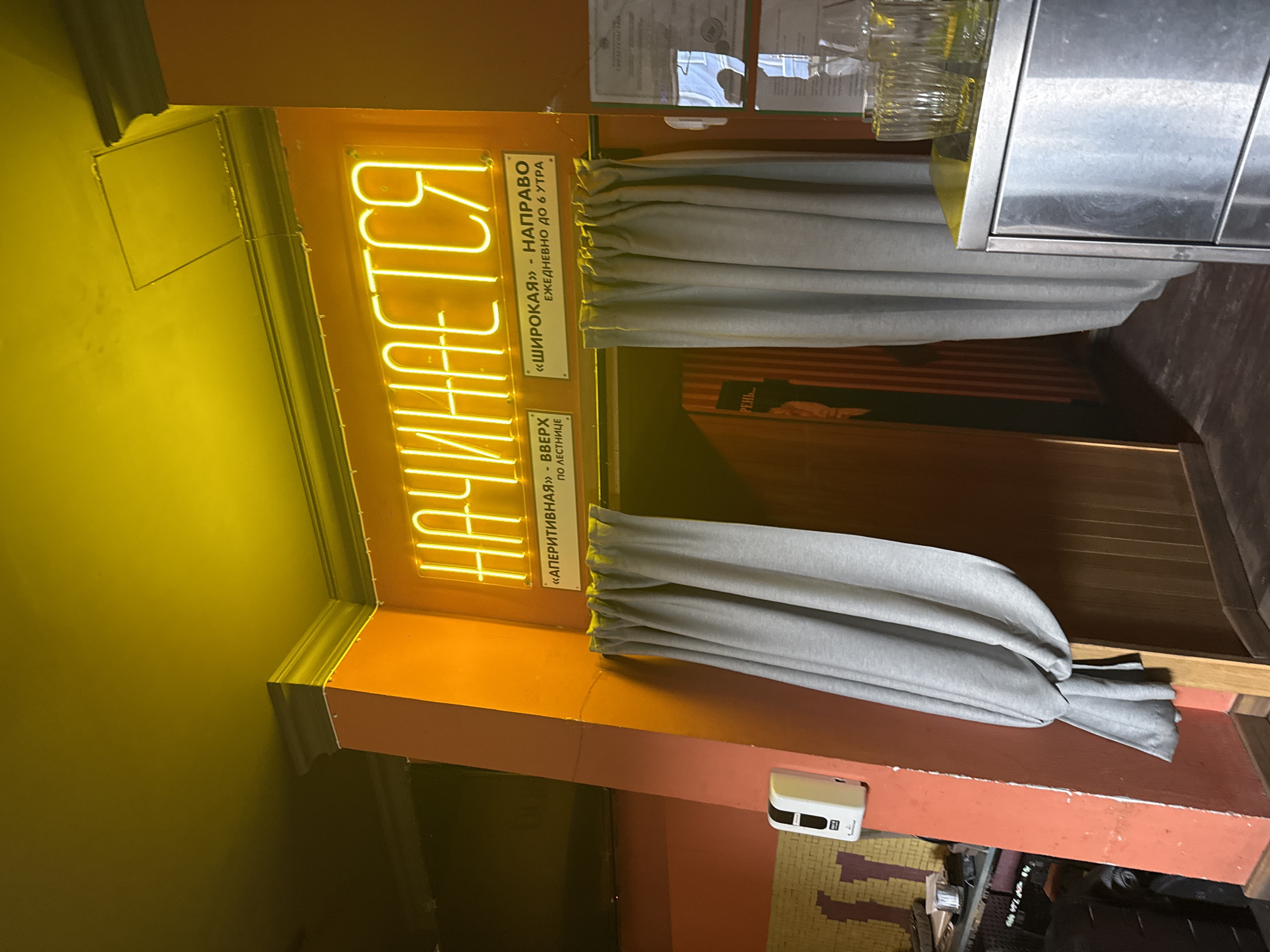 The image captures a scene inside a Moscow establishment. A neon sign spelling "НАЧИНАЕТСЯ" in Cyrillic is illuminated in a bright, yellowish light. Underneath the sign are two plaques with Cyrillic text: "«АПЕРИТИВНАЯ» - ВВЕРХ ПО ЛЕСТНИЦЕ" (Aperitif - Up the stairs) and "«ШИРОКАЯ» - НАПРАВО ЕЖЕДНЕВНО ДО 6 УТРА" (Wide - Right, Daily until 6 AM). Two draped gray curtains flank the signage. A stainless-steel structure, possibly part of a bar or counter, appears on the right. A white electronic device is visible on the wall below the sign and curtains. The background shows hints of an interior space with wooden accents.